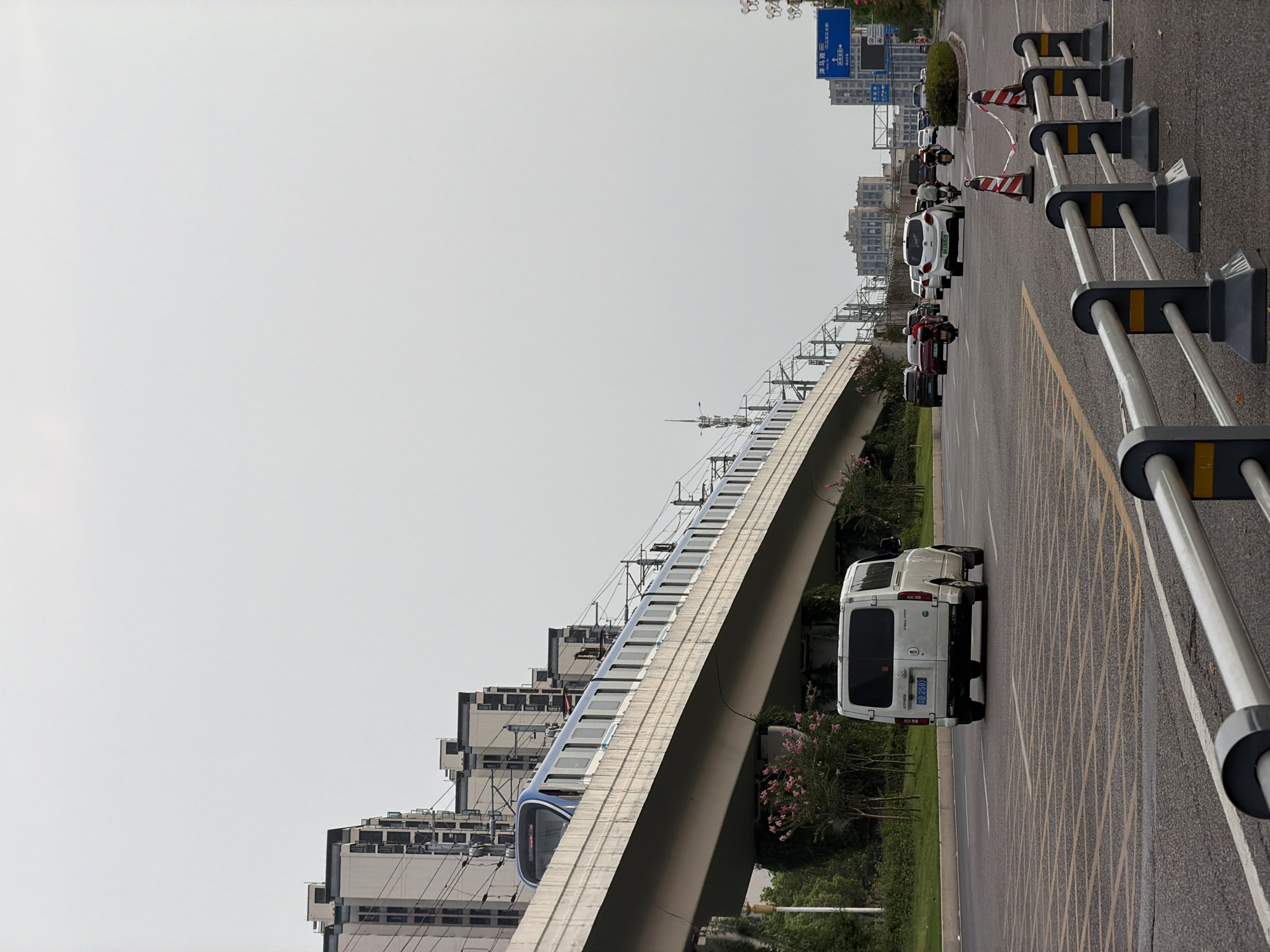 The image depicts an elevated metro train on a concrete track passing over a street in Jiangjin, China. The metro train is blue and white with multiple carriages. Below the track, on the street, there are several vehicles including a white van, cars, and motorcycles. The street has yellow and white lane markings. In the background, there are tall residential buildings and some street signs. The sky is overcast. The scene appears to be an urban environment with modern infrastructure.