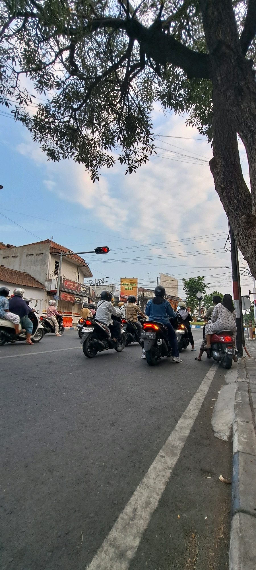 The image captures a street scene in Pasuruan, Indonesia, likely during daylight hours given the illuminated sky. Several individuals are riding motorcycles, indicating a common mode of transportation in the area. The road appears to be paved, with a white line marking a lane. On the right side of the road, a sidewalk with a concrete curb is visible, bordering a grassy area and a tree. Buildings line the left side of the street, some of which appear to be shops or residences, with signage in Indonesian. A traffic light with a red signal is prominently displayed, suggesting traffic is halted. Power lines are strung across the sky, a typical urban feature. The presence of motorcycles, the style of the buildings, and the signage all point to a location in Indonesia.