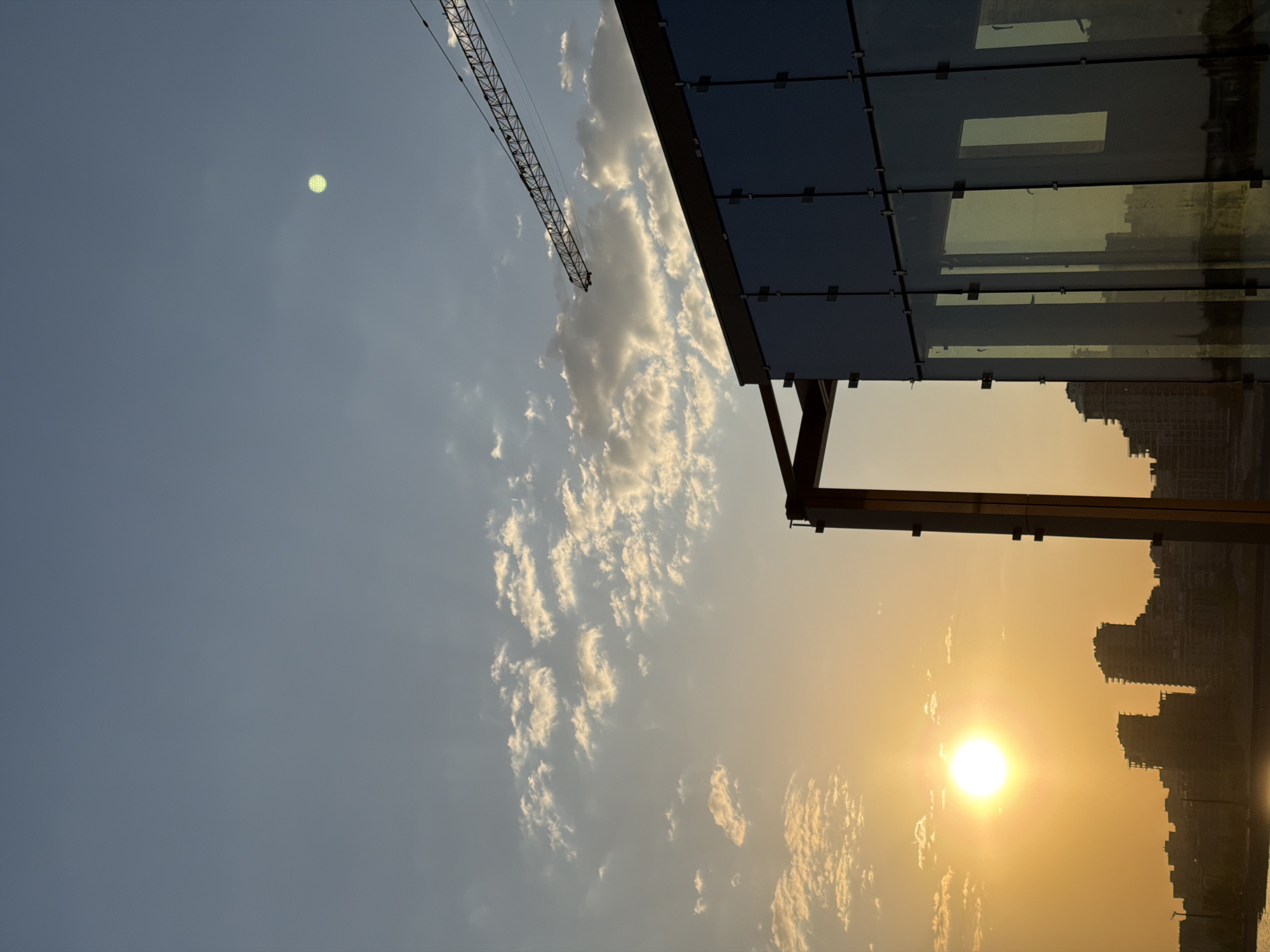 The image shows a view in Kazan, Russia at sunset. To the right, the corner of a modern building is visible with a glass facade reflecting the surrounding scene. The setting sun is positioned near the horizon line, casting a warm golden glow across the sky and illuminating the skyline of Kazan in silhouette. A construction crane is visible in the upper left of the frame against the blue sky and scattered clouds.