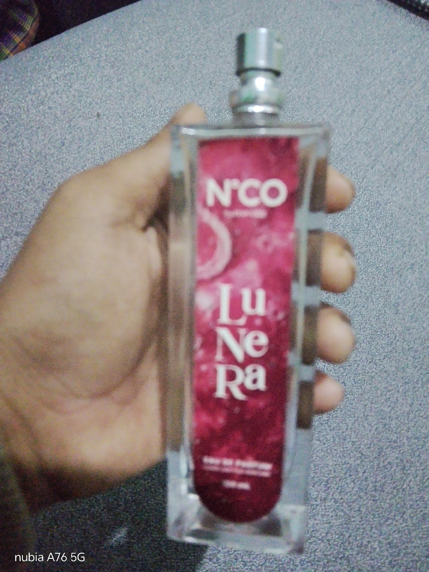 A hand holds a clear glass bottle of perfume. The bottle is rectangular with a silver cap and a pink label that has white text on it. The text on the label reads "N'CO," "Lu Ne Ra," and then some smaller text below that is illegible. The background is a grey speckled surface. The image is a close-up of the perfume bottle and the hand holding it. The location is Binuang, Indonesia.
