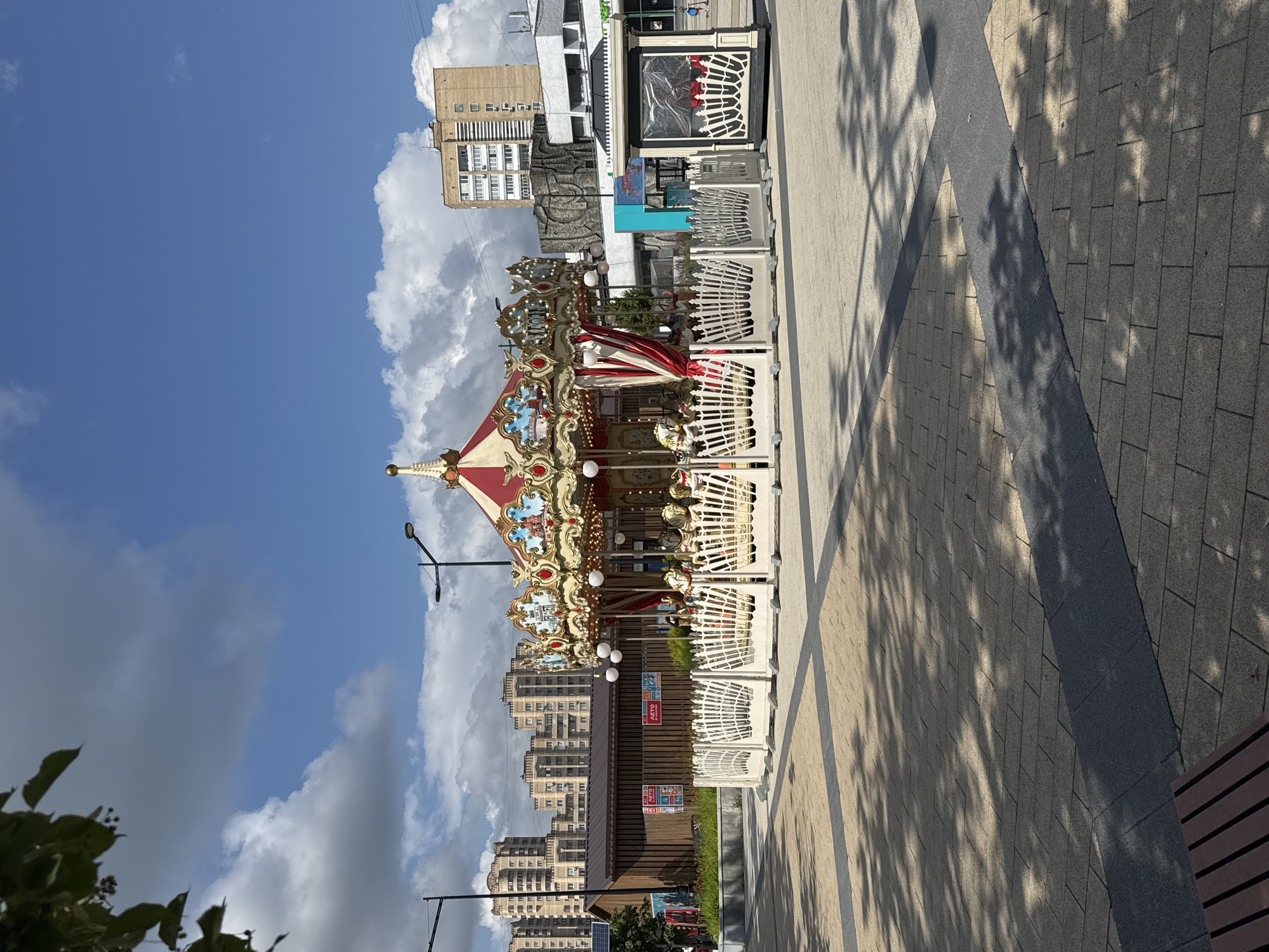 In Zelenograd, Russia, on a sunny day with scattered clouds, a bright red and gold carousel with white fencing stands in a paved outdoor plaza. The carousel features ornate decorations and several white horses, some of which are visible. Behind the carousel, a long, low building with a dark brown wooden facade is present, with signs on it. Further in the background, apartment buildings of various heights and architectural styles are visible, some appearing to be modern residential complexes with light-colored facades and numerous windows. Streetlights are positioned around the plaza. The ground is paved with a pattern of light and dark square tiles, with shadows of trees cast across it. A small, enclosed booth with a dark roof and transparent walls is situated to the right of the carousel.