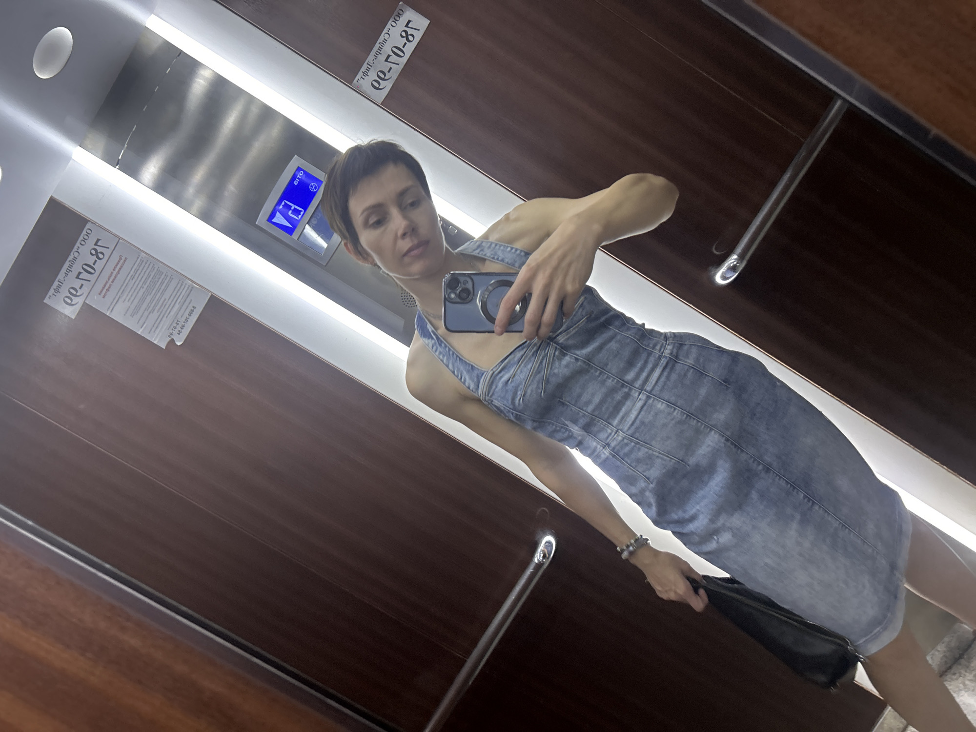 In the city of Kemerovo, Russia, a woman stands in an elevator with wooden paneling and stainless steel accents. She is taking a mirror selfie with her smartphone, which is held in her right hand. The woman is wearing a denim halter dress and a bracelet on her left wrist. She is also carrying a black handbag in her left hand. The elevator has a digital display showing the number "73" and a circular indicator with a face. A sign with Cyrillic text is affixed to the elevator wall.