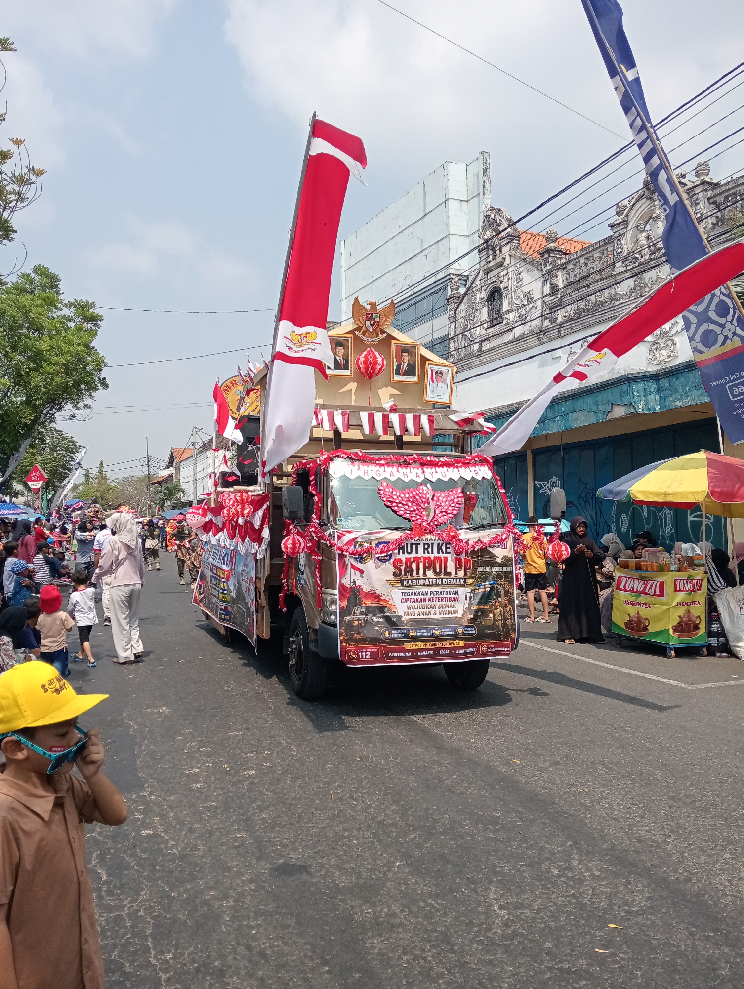 This is a photograph taken from a street-level perspective, looking towards a parade procession in the city of Demak, Indonesia. The central subject is a decorated truck, likely part of a Satpol PP (Public Order Agency) event, adorned with red and white banners and flags, and featuring portraits of national figures. The truck is moving down a paved street lined with buildings, trees, and a crowd of onlookers.
In the foreground, a young boy wearing a yellow cap and brown shirt is observing the parade, holding his sunglasses. The street is asphalt and appears to be a public road with some wear and tear. On the right side of the street, a vendor cart with colorful umbrellas is selling beverages, with customers gathered around.
The buildings in the background exhibit traditional Indonesian architecture, with ornate facades and tiled roofs. The sky is partly cloudy, indicating daylight. The overall scene depicts a lively public event, possibly a celebration or commemoration, with a strong sense of community participation. The presence of national flags and patriotic imagery suggests a connection to Indonesian national identity and local governance.