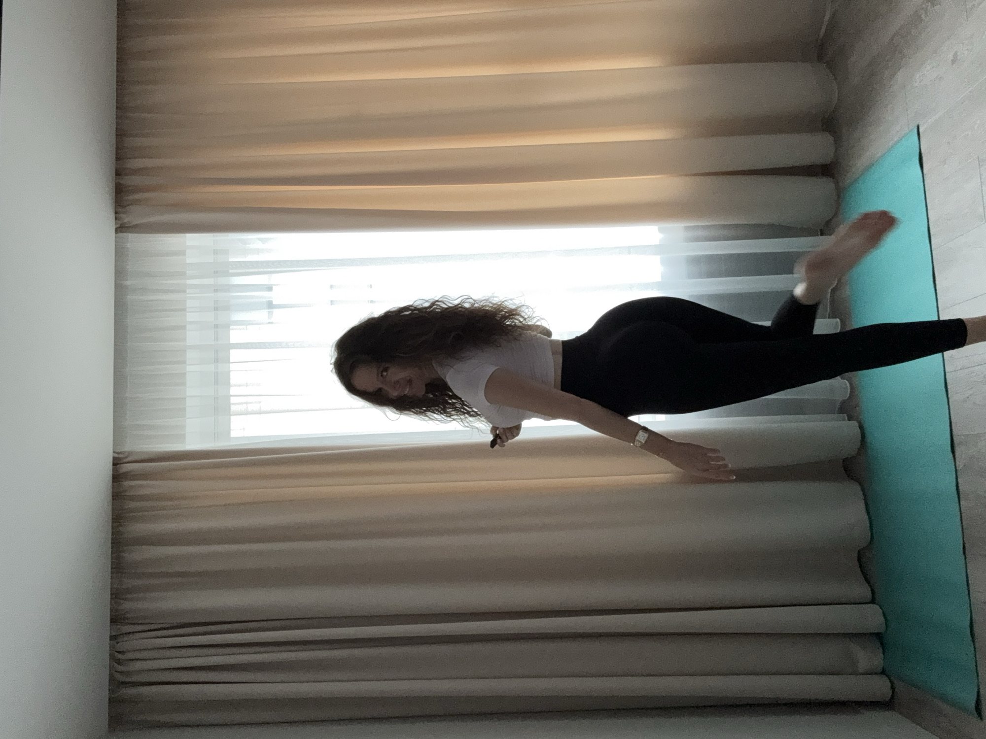 A woman with curly brown hair is standing on a turquoise yoga mat in front of light-colored curtains and a window. She is wearing a white crop top and black leggings. Her body is angled away from the camera, and she is looking over her shoulder with a smile. She is holding a black object in her right hand. A watch is visible on her left wrist. The room appears to be a residential interior, possibly a bedroom or living room, with light-colored walls and a wooden floor. The lighting suggests it is daytime, with natural light coming from the window. There is no visible indication that this location is in Moscow, Russia.