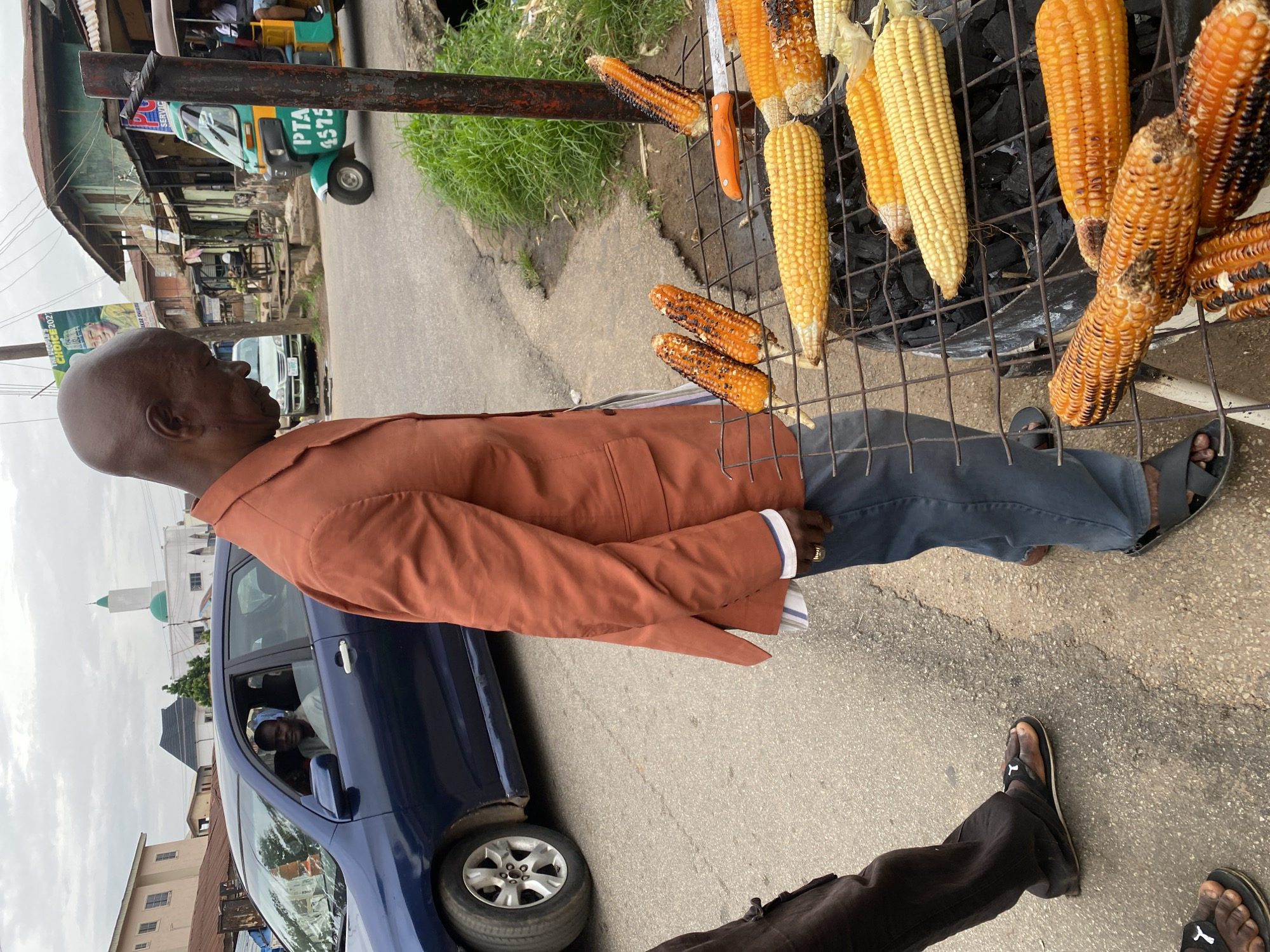 In Jos, Nigeria, a man in an orange blazer and blue pants stands over a charcoal grill laden with roasting corn. The man, with a shaved head, looks down at the corn. The grill, made of wire mesh, is positioned on a street. Several ears of corn are visible, some partially charred, others golden yellow. A white and orange knife rests on the grill. Behind the man, a blue car is parked, and its driver can be seen in the driver's seat. In the background, buildings, a white mosque with a green dome, and a colorful auto-rickshaw are present, suggesting a busy urban environment. The sky is overcast.