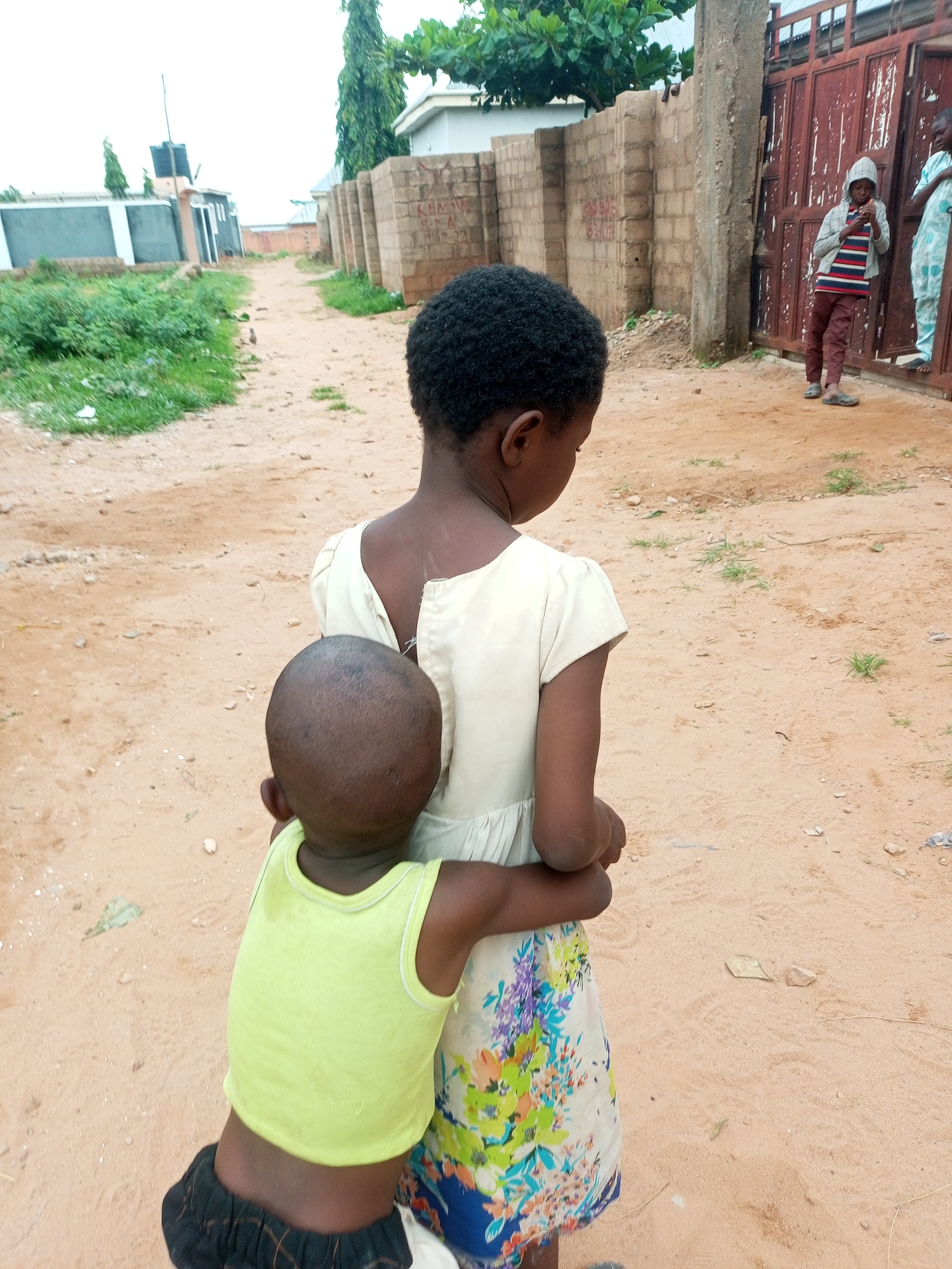 In Tempure, Nigeria, a young child wearing a lime green tank top and black checkered shorts embraces a slightly older child from behind. The older child, seen from the back, wears a cream-colored dress with a floral pattern. They stand on a dirt path in what appears to be a residential area, indicated by brick walls and buildings in the background. To the right, a person in a hooded gray jacket with striped accents and brown pants stands near a red metal gate, and another person in a light blue garment is visible further to the right. The environment is dusty with sparse vegetation.