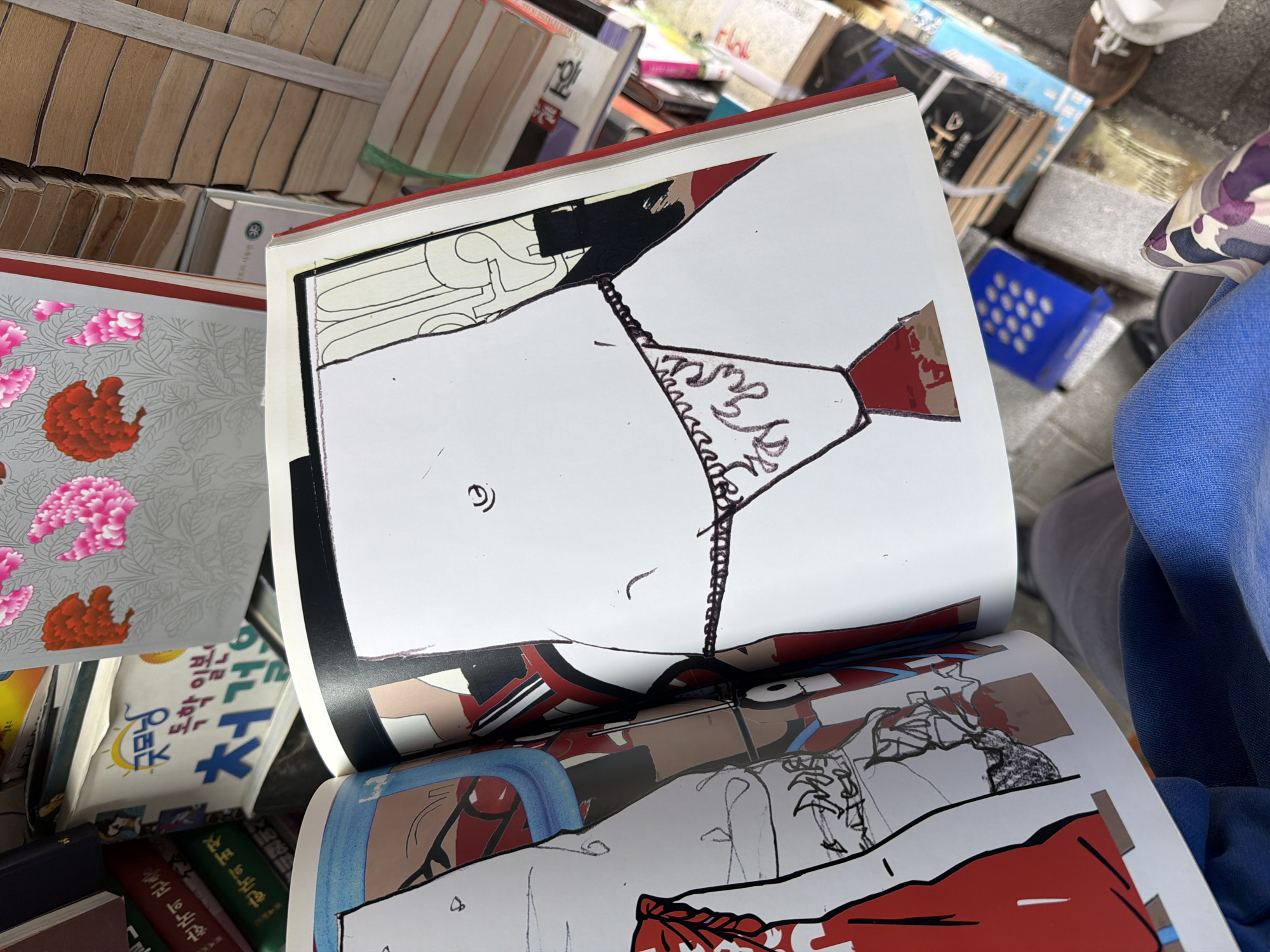The image shows a close-up of an open book resting among a stack of other books and objects. The book displays cartoon-style illustrations of nude or semi-nude figures. A person wearing a blue garment is visible to the right of the book. Other books in Korean are also visible. The environment suggests a possible book stall or second-hand book market. This is happening in Seoul, South Korea.