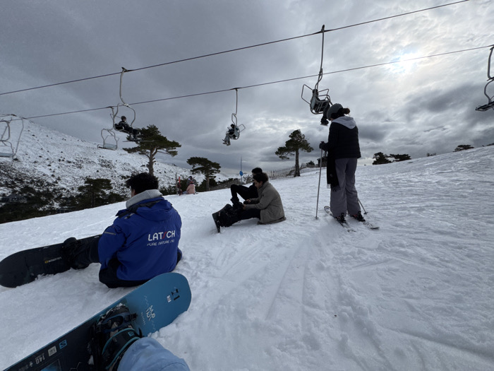 A snowy mountain scene shows a group of people resting on the slopes. A person in a blue jacket sits with a snowboard, while others sit nearby. A skier stands with poles. Overhead, ski lift chairs ascend the mountain, some carrying passengers. The sky is cloudy and overcast, with a hint of sunlight breaking through. Scattered trees dot the landscape. The scene conveys a winter recreational activity in a mountainous setting.