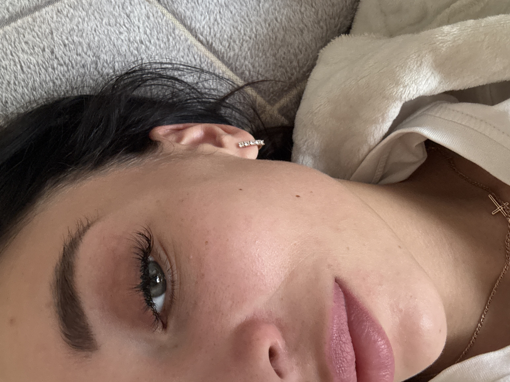 A close-up, slightly tilted, portrait-style shot of a light-skinned woman with dark hair. Her face is the primary focus, with one grey-green eye clearly visible, defined eyebrows, and pink lips. An earring with small stones is visible on her left ear. The woman is lying down on a grey textured surface, possibly a blanket or cushion. She is wearing a white top, and a gold necklace with a cross pendant is visible on her chest. The location in the city of Chaikovsky, Russia is not evident in the image.
