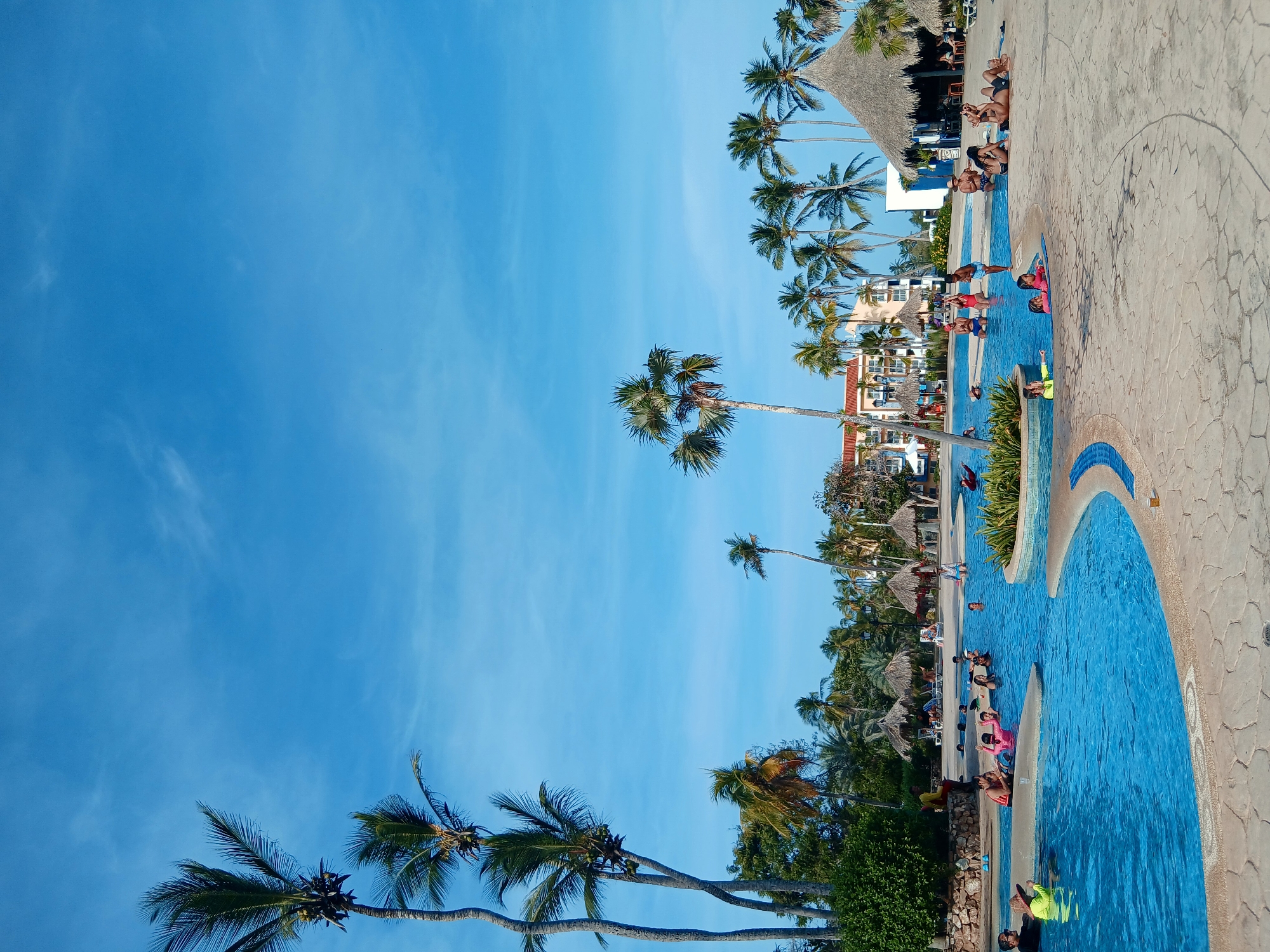 The image shows a view of a resort swimming pool in Playa El Agua, Venezuela. Palm trees line the pool area, and people are swimming, sunbathing, and lounging around the pool. The sky is blue. There are thatched umbrellas. A multistory hotel building is visible in the background.