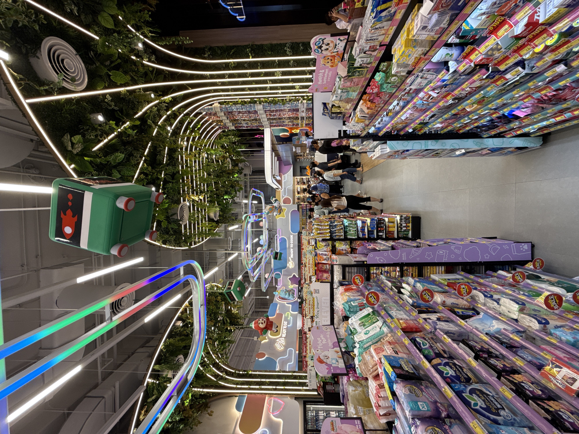 This image depicts the interior of a modern convenience store, possibly a 7-Eleven, located in Bang Kaeo Town Municipality, Thailand. The store features a vibrant and playful design, with colorful lighting, hanging decorative elements resembling a cartoon train, and an abundance of artificial greenery arching over the aisles.
In the foreground, multiple shelves are stocked with a wide variety of retail products. The shelves are organized and well-lit, with many items displaying price tags and promotional "SALE" stickers. Some of the visible products include what appear to be diapers, sanitary products, snacks, and beverages. The aisle in the foreground is relatively clear, suggesting it is either early in the day or not a peak shopping period.
Further into the store, several customers are browsing the merchandise. They are diverse in age and gender, dressed in casual attire. Interactions between customers are not clearly discernible, but they appear to be independently shopping.
The ceiling is a prominent feature, adorned with dynamic LED strip lighting in various colors, including blue, green, and pink, tracing curved pathways. The artificial greenery creates a naturalistic yet whimsical ambiance, contrasting with the typical sterile environment of many convenience stores. The overall impression is one of a retail space designed to be visually engaging and perhaps appeal to a younger demographic or families. The store is clean and well-maintained.