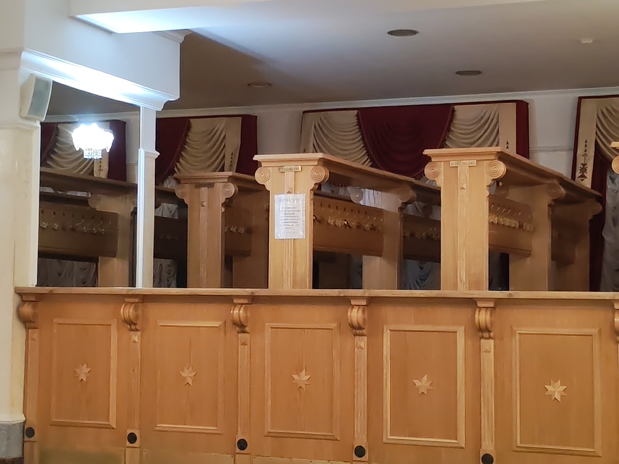The image displays the interior of an ornate cloakroom or checkroom, likely within a public building in Izhevsk, Russia. The scene is dominated by a long, light-colored wooden counter or barrier in the foreground, featuring decorative panels with carved star-like motifs. Behind this counter, a series of robust wooden columns with scrolled capitals rise, supporting an upper wooden structure.
Further back, sections equipped with numerous coat hooks are visible, indicating the facility's purpose. The ceiling is white and features recessed circular lighting. Additionally, a multi-bulb chandelier can be seen glowing in the distance on the left. The walls are decorated with a pattern of elaborate drapes in rich red and pleated beige/gold fabrics, contributing to a formal and classic aesthetic.
No people are present in the image, suggesting the area is empty. On a central wooden column, a white paper notice, which appears to be in Cyrillic, is attached, though its text is unreadable. Above this notice, a small inscription on the wooden structure reads "№167 240", possibly an identification or section number. A white rectangular speaker or vent is mounted on a column on the far left. The setting evokes a sense of traditional elegance and order, characteristic of a historical or grand public venue.