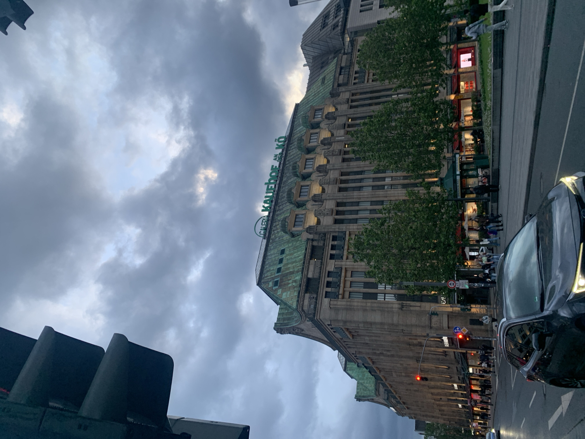 This detailed image captures an urban street scene at Königsallee in Düsseldorf, Germany, during dusk or early evening. The prominent central subject is the large Galeria Kaufhof department store, identifiable by the green "GALERIA KAUFHOF AN DER KÖ" sign on its rooftop. The building features a multi-tiered, green-patinated copper roof with numerous dormer windows and a grand, light-colored stone facade characterized by vertical pilasters and recessed windows.
The sky is largely covered with heavy, dark grey clouds, with patches of lighter sky and faint sunlight visible towards the right. A multi-lane street, appearing slightly wet and reflective, occupies the lower portion of the image. In the immediate foreground, a dark-colored SUV, possibly a Toyota RAV4, is angled slightly right, its headlights illuminated. On the left side of the street, multiple traffic lights display red, and other vehicles are visible further down.
Wide sidewalks line both sides of the street, separated from the road by areas planted with mature deciduous trees with green foliage. Numerous pedestrians are present on the sidewalks, particularly on the right side in front of the department store, engaged in walking. The ground floor of the Galeria Kaufhof building features many brightly lit shop windows, casting a warm glow on the street level and indicating retail activity. The overall lighting suggests twilight conditions, with artificial lights complementing the dwindling natural light.