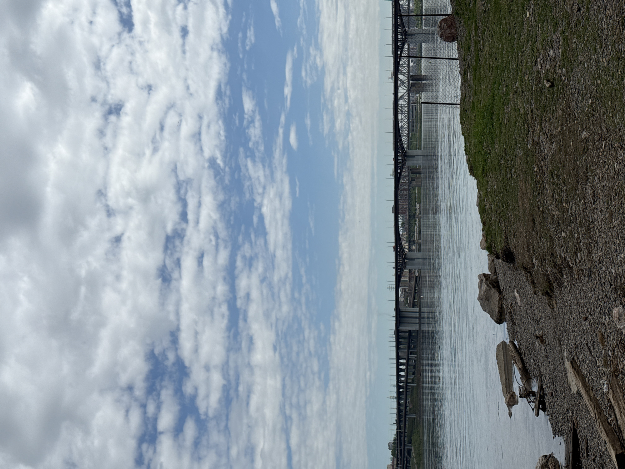 The image depicts the Yenisey River with a bridge in Krasnoyarsk, Russia. The sky is predominantly blue with many white clouds. The bridge is a steel structure, running horizontally across the frame. On the right side of the image, there is a gravelly shoreline with some greenery. The water is relatively calm with slight ripples. The city skyline is visible in the distance, below the bridge.