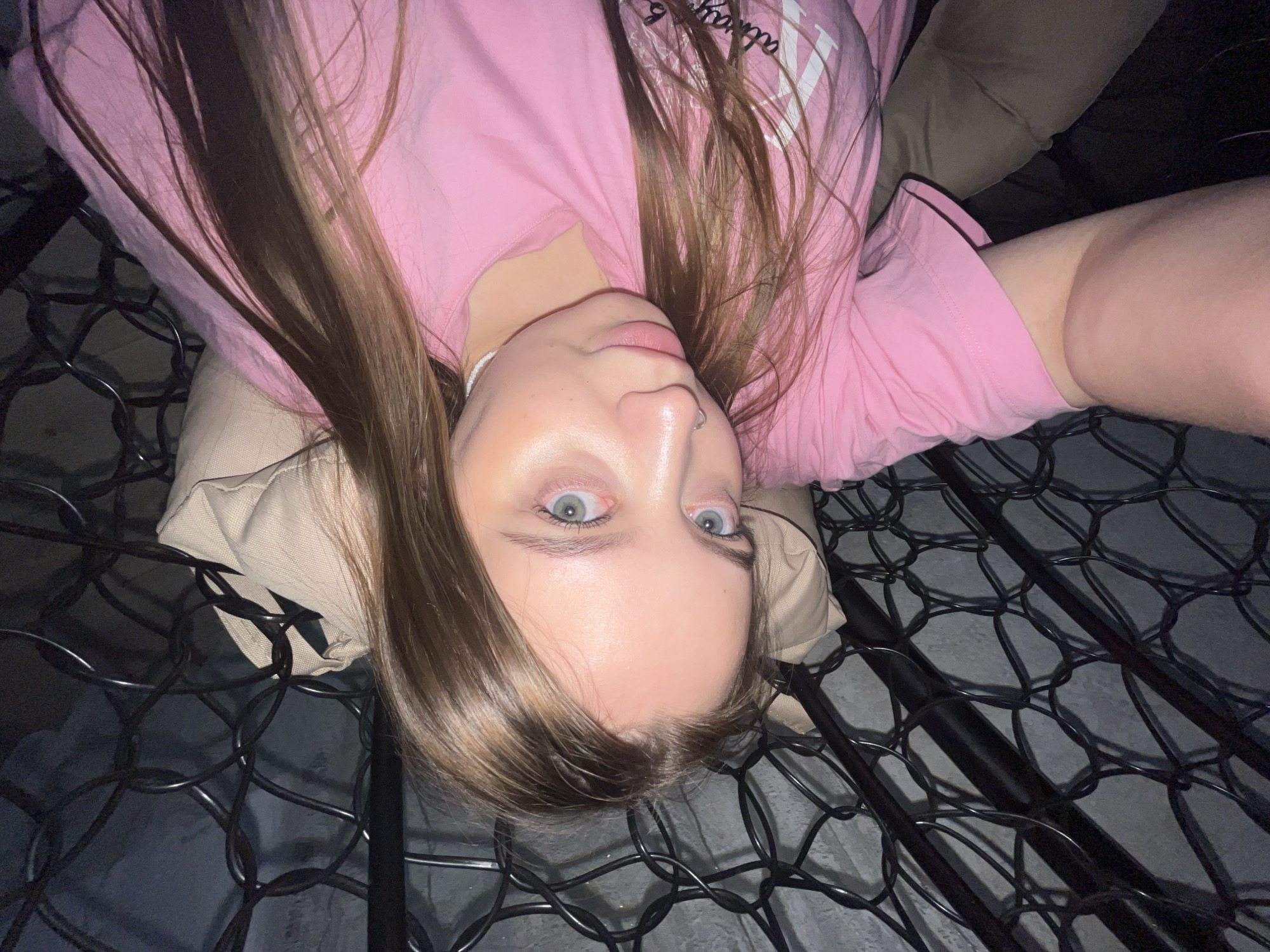 A young woman is positioned upside down, with her face centered in the frame. She has light skin, light eyes, and shoulder-length light brown hair. She is wearing a pink t-shirt. The background shows what appears to be a black metal frame, possibly a bed or cot, with a grey surface beneath it. The photograph was taken indoors, location of Novopokrovskaya, Russia is undetermined.