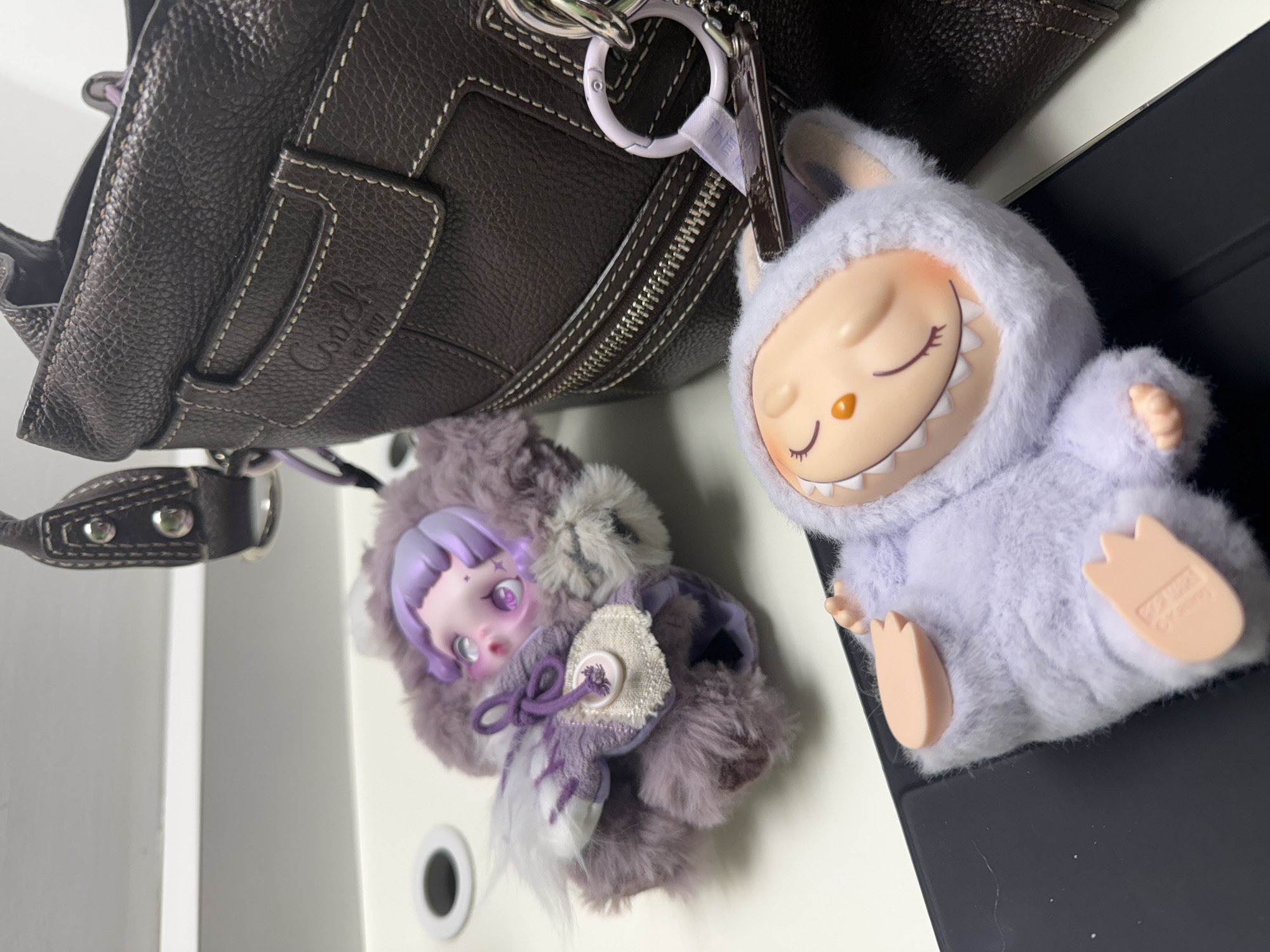 A close-up shot shows a brown Coach purse, oriented diagonally, with two plush toys attached to it. One is a furry, purple doll-like figure with a pale face, purple hair, and a buttoned cloak. The other is a light purple, fuzzy monster-like toy with a wide grin and pale pink hands and feet. The background includes a white surface and a dark, possibly electronic, device. The location, Yerevan, Armenia, cannot be determined from the image alone.