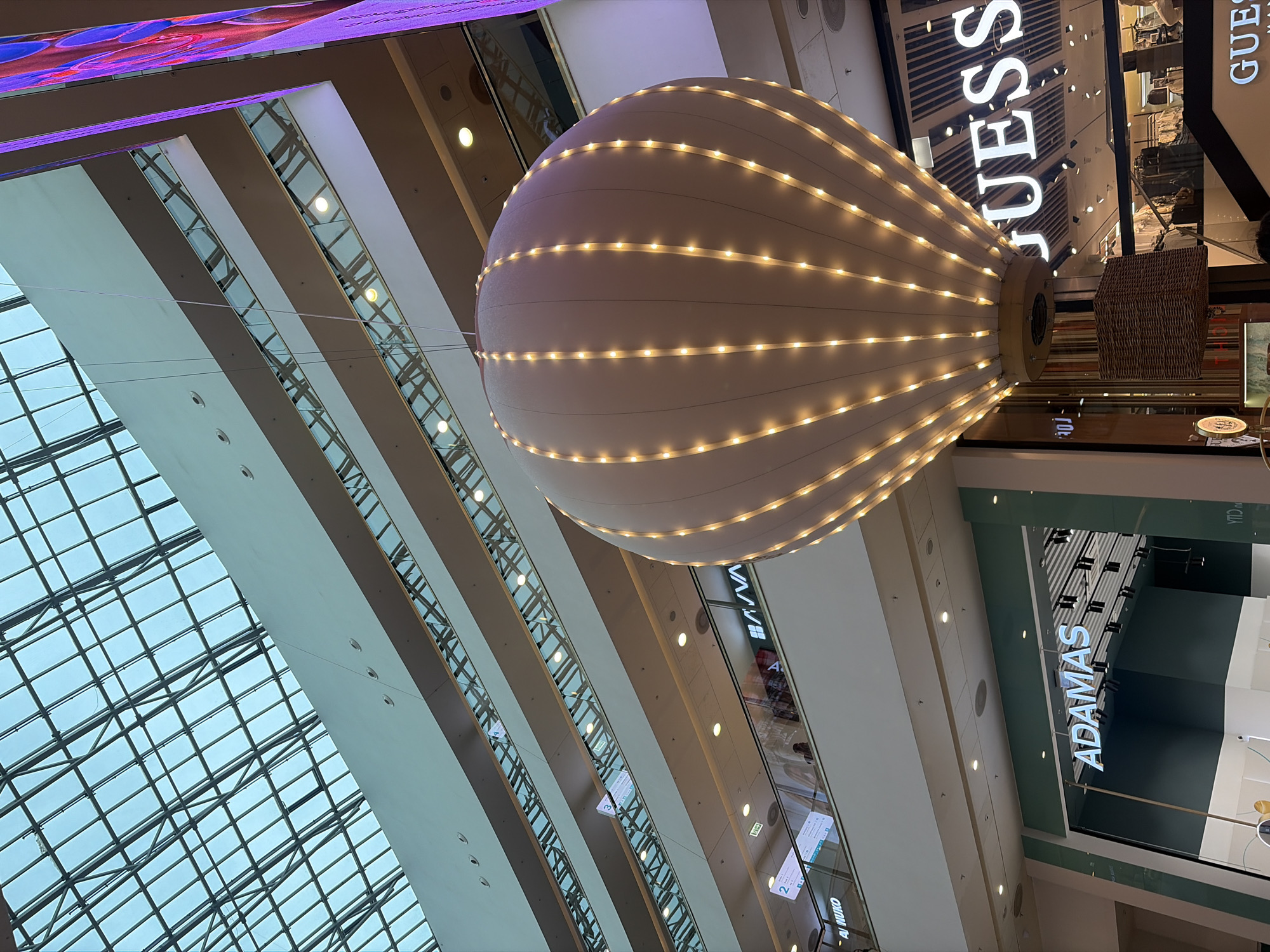 The image shows the interior of a multi-story shopping mall. A large, illuminated hot air balloon-shaped decoration hangs from the ceiling, with a wicker basket beneath it. The mall features multiple levels with escalators, glass railings, and various storefronts, including "ADAMAS" and "GUESS." The ceiling is a glass and metal structure that allows natural light to filter in. The overall impression is a modern, well-lit retail environment. The specific location in Saint Petersburg, Russia is not indicated in the image. There are a few individuals visible on the escalators and walkways.