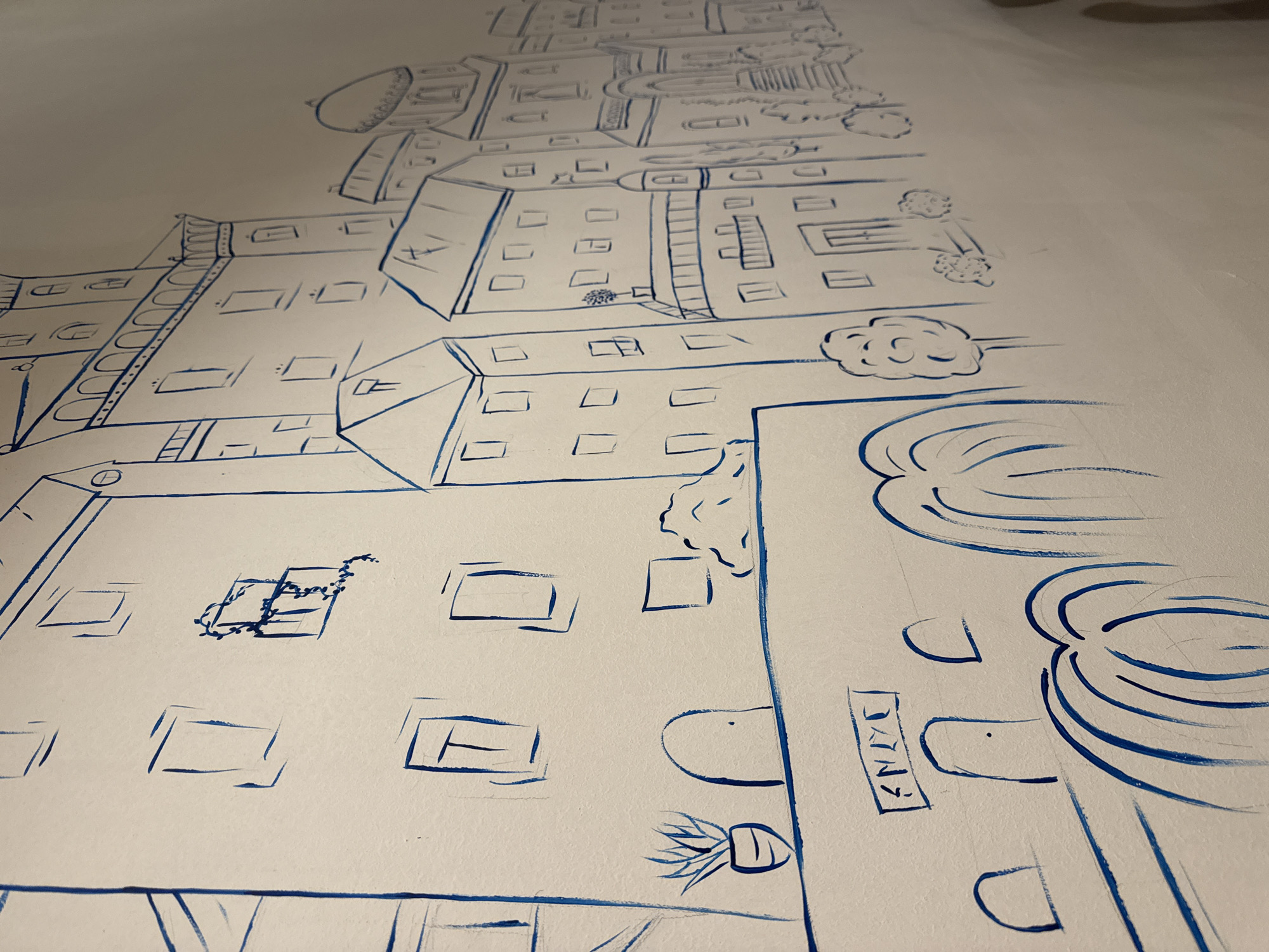 The image is a hand-drawn, blue-ink sketch of a cityscape in Izhevsk, Russia. The perspective is from an elevated viewpoint, looking down on the rooftops and architectural details.
Buildings: The drawing depicts various buildings, including structures with flat roofs, gabled roofs, and a building with a dome. Windows are represented as simple squares or rectangles.
Street: A street is shown running through the drawing.
Landscaping: Some trees and bushes are included as simplified cloud-like forms.
Details: The drawing includes details like a ladder on the side of a building, and a decorative design of climbing foliage or vines on the roof of one building. A rectangular sign with text is visible at the bottom right.
