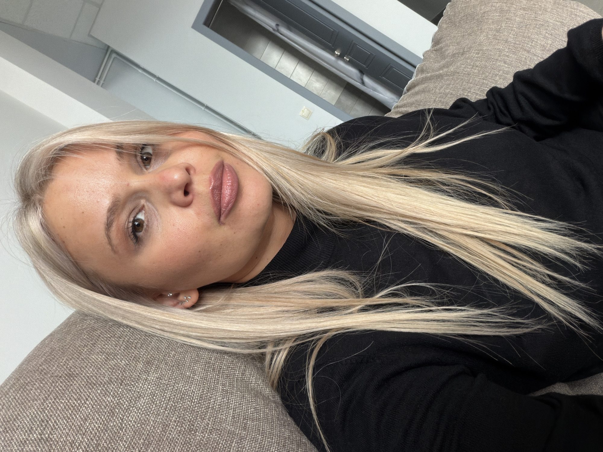 A woman with light blonde hair is lying on a beige sofa. She is wearing a black top. The background includes a gray door and a white ceiling. The location is presumably indoors, possibly in Sarapul, Russia.