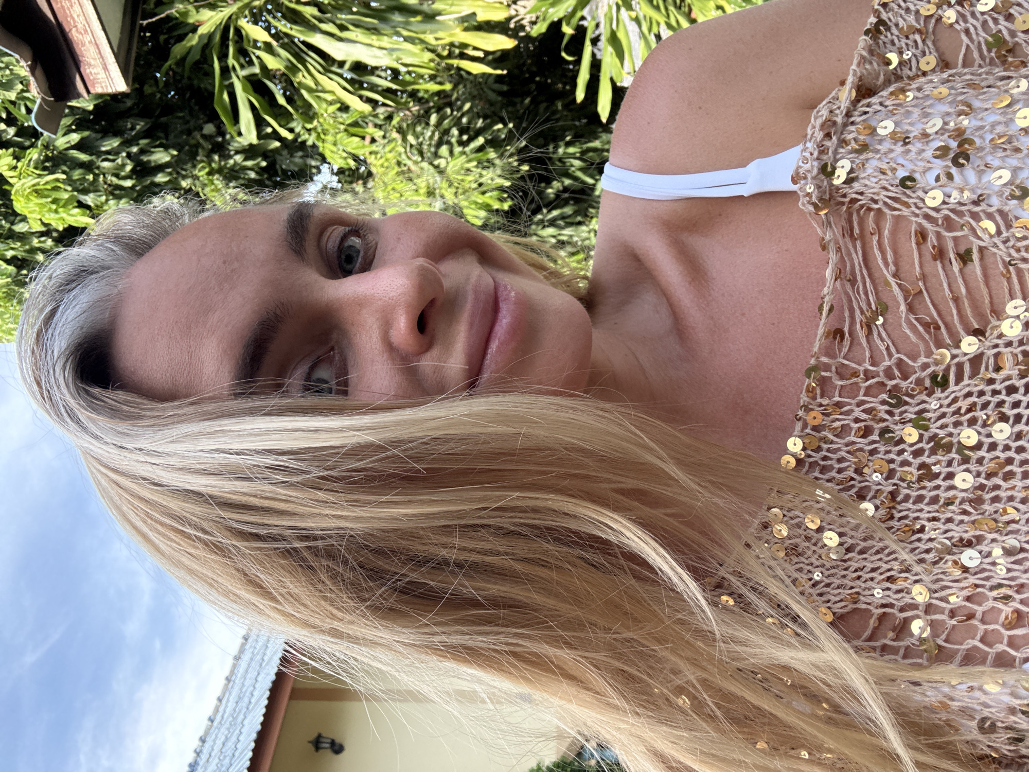 A medium shot captures a Caucasian woman with blonde hair and blue eyes outdoors, possibly in the Ko Samui District of Thailand. She is looking towards the camera with a slight smile. Her hair is long and appears windswept, with some strands falling across her face. She is wearing a white top with thin straps, and an open-knit, beige cover-up adorned with gold sequins. In the background, lush green foliage and a portion of a building with a metallic roof are visible. The sky above is a light blue with scattered clouds.