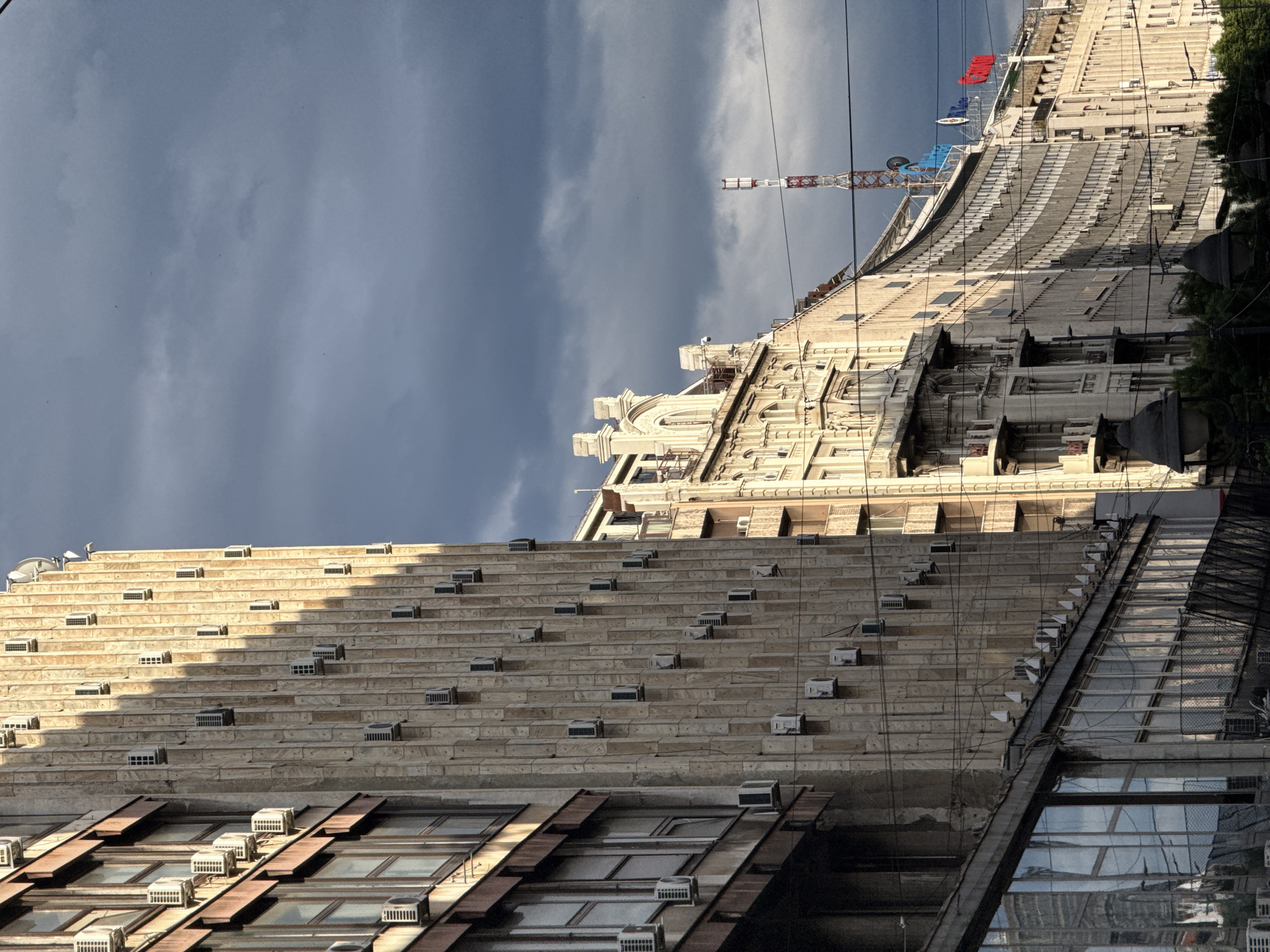 The image depicts two buildings in Belgrade, Serbia, under a cloudy sky. The foreground building has a facade composed of rectangular brick-like segments, each equipped with a small air conditioning unit. Below this, there are windows with metal awnings, some also equipped with air conditioning units. The background building is taller, with ornate architectural details and windows. A construction crane is visible behind this building. Power lines stretch across the upper portion of the image.