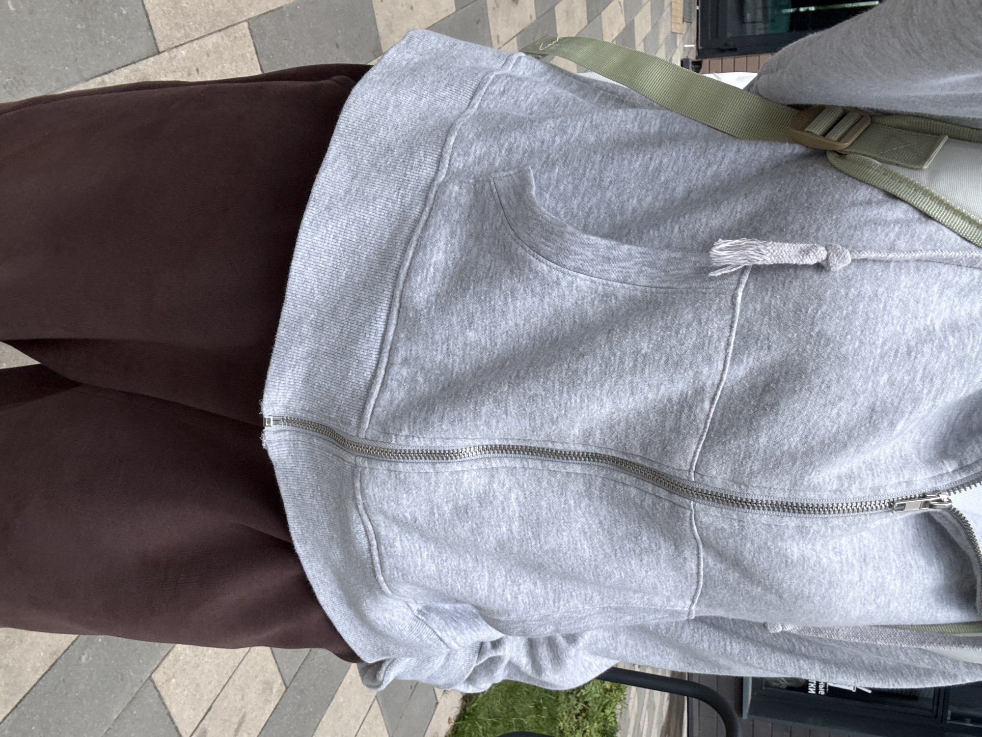The image shows a close-up view of a person wearing a light gray zip-up hoodie and dark brown sweatpants. A pale green strap from a backpack or bag is visible over the person's left shoulder. The person is standing on a sidewalk paved with light and dark gray rectangular tiles. In the background, a building with dark windows and some greenery is visible. On the building, text in Cyrillic characters can be partially seen. The scene is outdoors, suggesting it is a public space within the city of Lyubertsy, Russia, given the context. The focus is on the clothing and the immediate surroundings of the person.