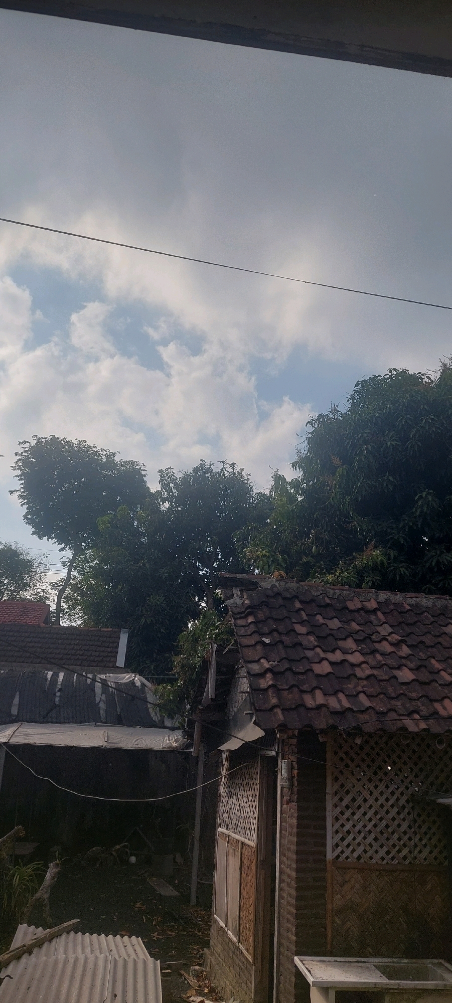 The image captures a somewhat overcast sky with scattered clouds above a residential area. In the foreground, there are rooftops of houses constructed with terracotta tiles and corrugated metal. Lush green trees dominate the midground, with their dense foliage creating a stark contrast against the sky. In the foreground, a building with a lattice-like wall and wooden panels is visible, suggesting traditional architecture. The scene appears to be in a rural or semi-urban setting within Lumajang, Indonesia, characterized by modest dwellings and abundant vegetation. There are no people visible in the frame.