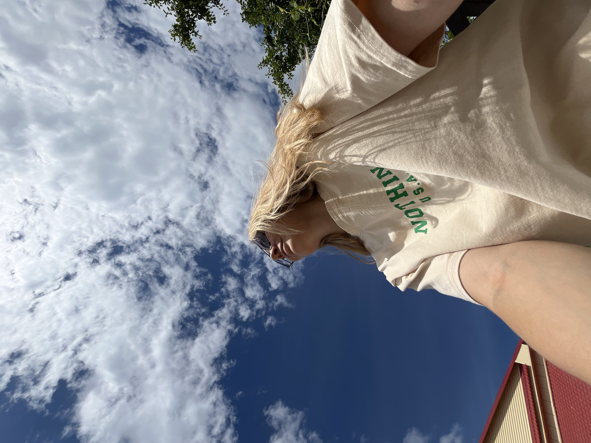 The image shows a low-angle shot of a young woman with blonde hair and sunglasses, looking down. She is wearing a light-colored t-shirt with green lettering that appears to say "NOTHIN U.S.A." in a circular pattern on the front. The background is a bright blue sky with scattered white clouds, suggesting a sunny day. Some green foliage is visible at the top of the frame, and a portion of a building with a red roof is in the lower right corner. The perspective suggests the photo was taken outdoors, possibly at midday given the bright lighting. The woman's expression is neutral, and the overall mood is casual and relaxed. The location context indicates this image was taken in Chaikovsky, Russia.