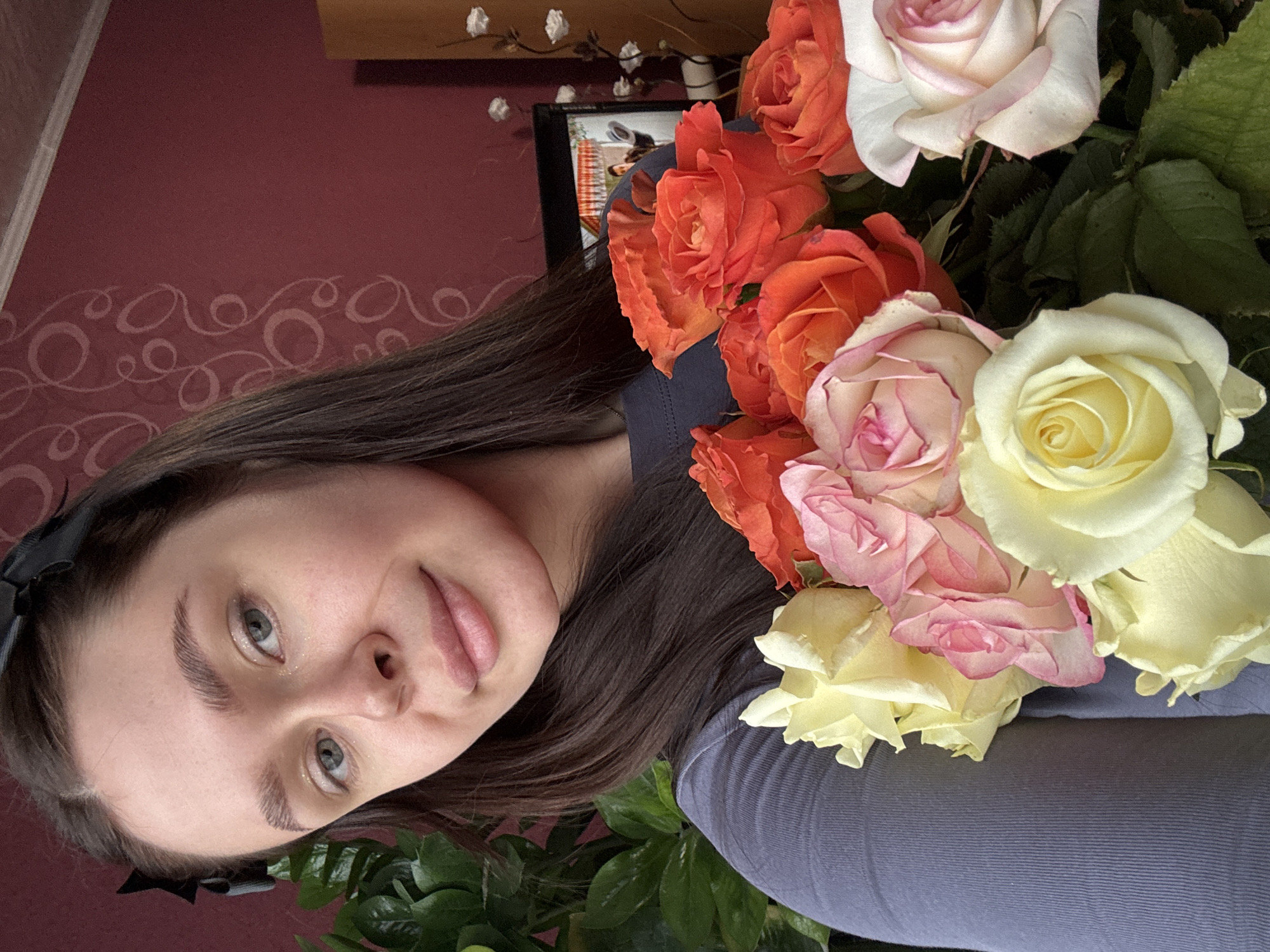 A young woman poses with a bouquet of roses. The roses are various colors, including orange, white, pink, and yellow. The woman is fair-skinned with light eyes and dark hair. She is wearing a dark-colored shirt and a black bow in her hair. Behind her is a red wall with a pattern, a television, a wooden cabinet, and artificial flowers. The location is unknown and the city of Dubrova, Russia cannot be confirmed.
