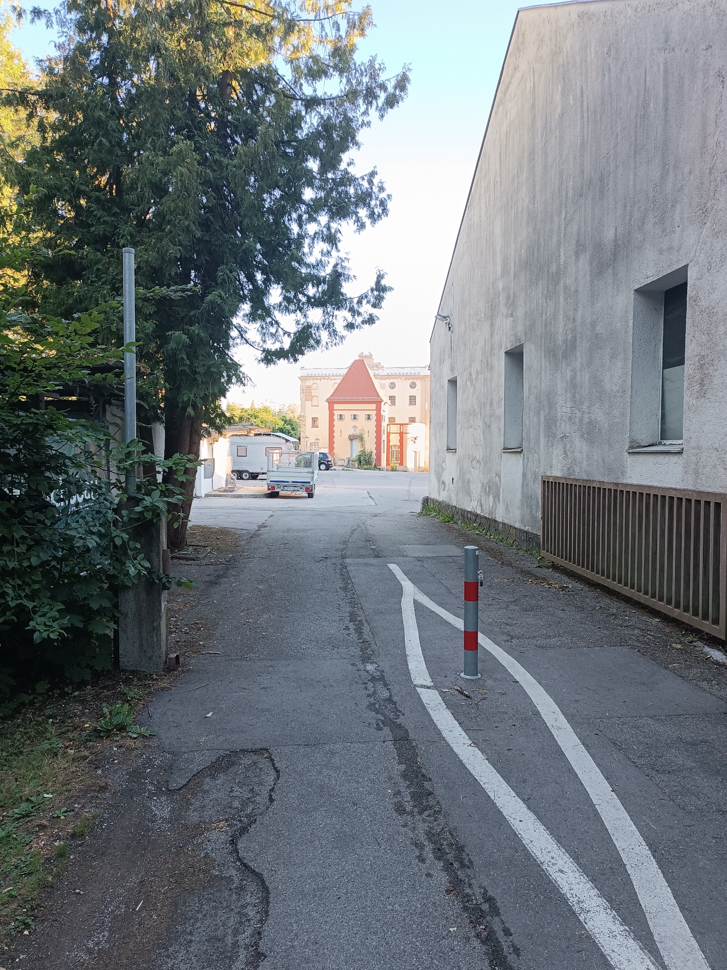 This image depicts an asphalt road in Fürstenfeldbruck, Germany, leading towards a large, multi-story building with a red roof and pale yellow facade. The road is flanked by a tall, light-colored building on the right with a metal fence along its base, and on the left, by a large tree and dense foliage. The road itself has white lane markings and a red and grey striped bollard. In the background, a small trailer is parked. The overall scene suggests a street or pathway within an industrial or institutional complex.