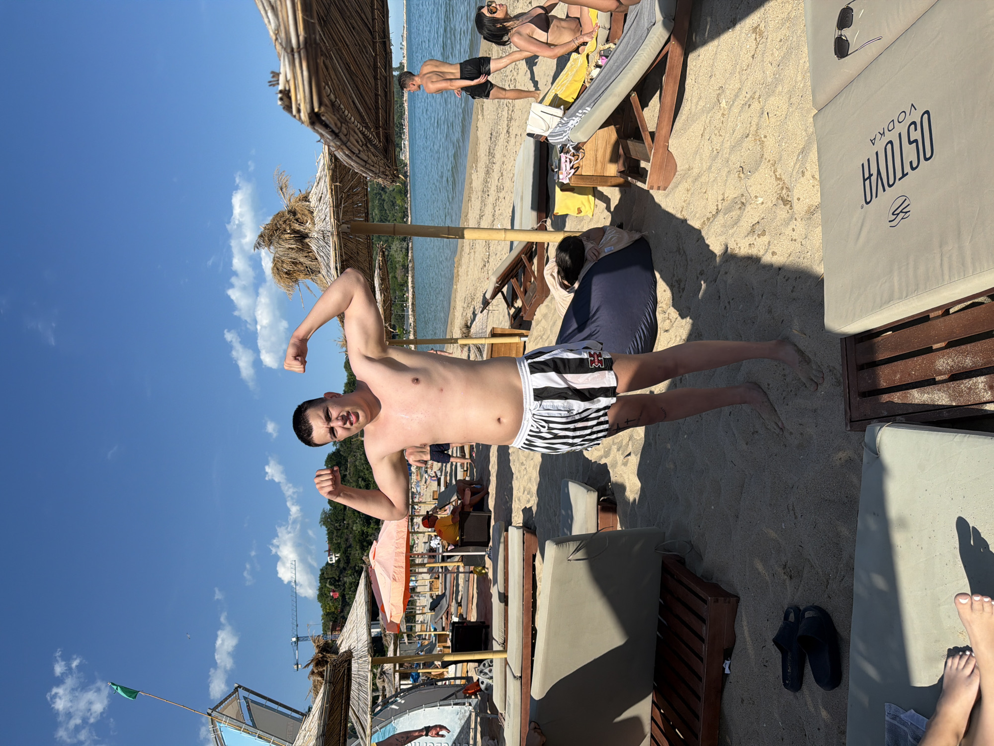 The image depicts a sunny day at a beach in Varna, Bulgaria. In the foreground, a young man with dark hair, wearing black and white striped shorts, is posing by flexing his biceps. He stands on the sandy beach, which is lined with beach chairs and thatched umbrellas. In the background, the sea is visible, with a few people scattered along the shoreline and some structures and hills in the distance. The overall scene is a typical beach environment on a clear, bright day.