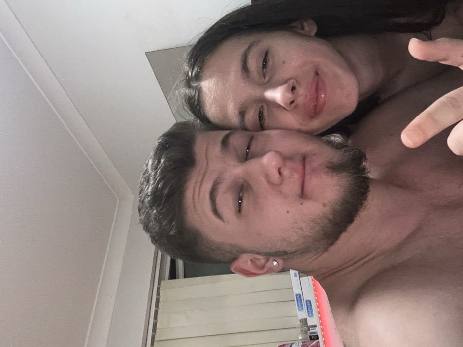 The image depicts two individuals, one male and one female, in an indoor setting. The male, positioned on the left, has light skin, short dark hair, a short beard and goatee, and a stud earring in his left ear. He appears shirtless, with his bare chest and shoulders visible, and is looking directly at the camera with a subtle expression. His left hand is partially visible in the lower right foreground, forming a V-sign with his index and middle fingers. The female, positioned on the right, also has light skin and long dark hair. Her head is close to the male's, and she appears to be leaning against him, looking directly at the camera. Her lips are full and appear glossy or slightly swollen. Her bare right shoulder is visible. Both individuals appear to be participating in a close-up photograph, likely a selfie.
The background features a white wall and white ceiling with simple coving. To the left, a window with vertical blinds is partially visible. Below the window, several stacked rectangular packages or boxes are present, with the word "KARELIA" visible on some. A red LED light strip casts a distinct red glow beneath these packages. To the right, behind the female, a rectangular mirror is mounted on the wall. The lighting is artificial and somewhat dim. The specific location within Corby, United Kingdom, cannot be determined from the visual evidence present in the image.