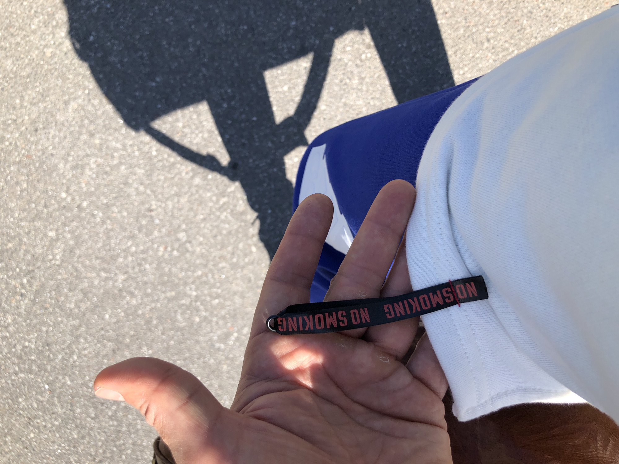 The image shows a close-up of a person's hand holding a black fabric tag with red text that reads "NO SMOKING". The tag is attached to a white garment, possibly a t-shirt, which is partially visible. The background is an outdoor scene with a gray, textured surface, likely asphalt or pavement, with sunlight casting a shadow of a person onto it. The person's hand is resting on blue fabric, likely pants or shorts. The overall lighting suggests it is daytime. The image does not provide any specific details that would allow for identification of the location within the city of Saint Petersburg, Russia.