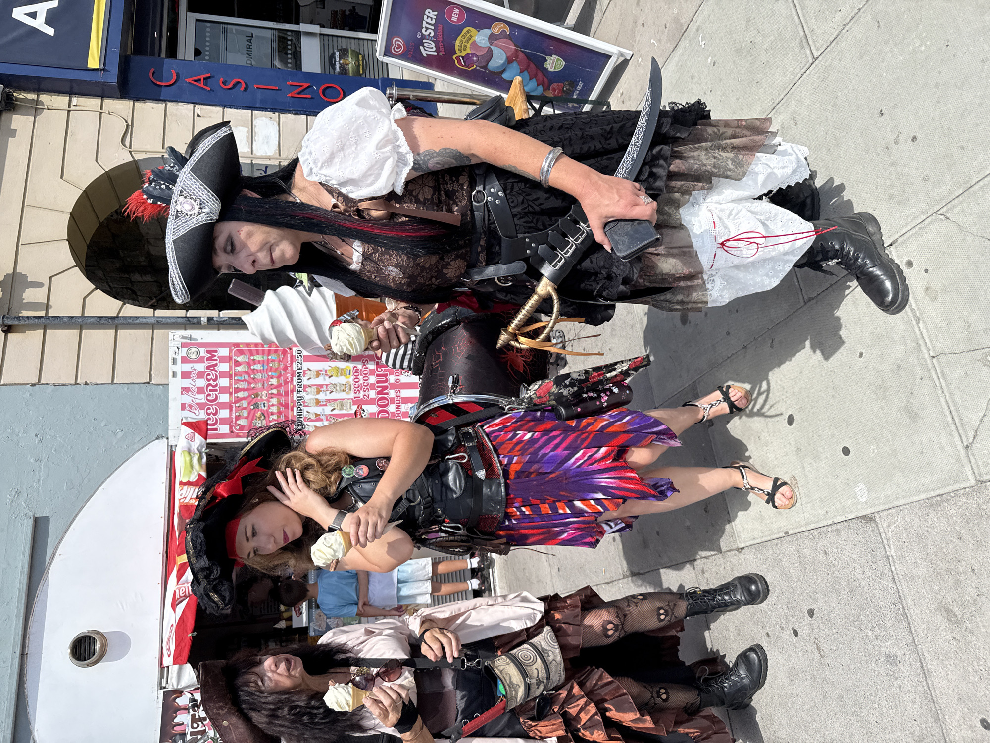 Three individuals dressed in pirate-themed attire are standing on a sidewalk in front of an ice cream shop. The person on the left, wearing a brown pirate hat, a light pink blouse, and a ruffled brown skirt with patterned fishnet stockings, holds a soft-serve ice cream cone. The middle person, with long brown hair and a red bandana, wears a black leather vest over a colorful, layered skirt and also holds an ice cream cone. They are both looking towards the left. The person on the right, with long black hair and streaks of red, sports a large, ornate pirate hat, a white puff-sleeved top, a brown patterned dress, a black leather belt with a sword attached, and a black skirt. They are holding an ice cream cone and looking towards the left, holding a knife in their right hand. A young person in a blue t-shirt and light-colored shorts is visible in the background, near the ice cream shop. An ice cream menu board is to the right, displaying various flavors and prices. The scene is illuminated by bright sunlight, casting shadows on the sidewalk. The background includes architectural elements of buildings, suggesting an urban or seaside location. The image appears to be taken on Marina, St Leonards-on-Sea, East Sussex, United Kingdom.