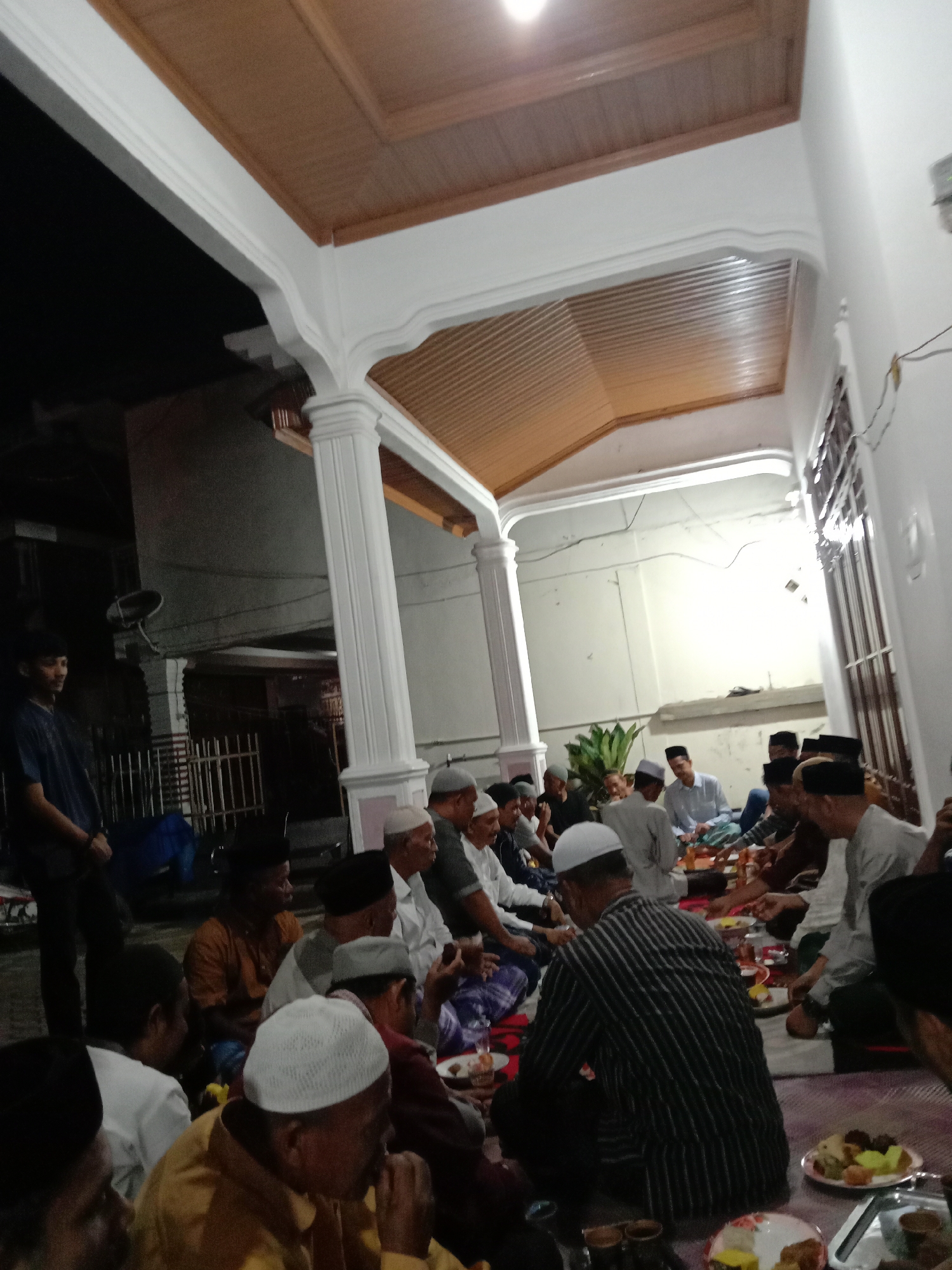 A gathering of people, mostly men, are sitting on a rug in an outdoor or semi-outdoor setting. Most of the men are wearing white or dark caps. Some are eating from plates. The setting appears to be a covered patio or outdoor extension of a building, supported by white columns. The ceiling above is made of brown wood panels. In the background, there's a fence and possibly another building. A young man is standing near the fence. Based on visual cues, this could be a social or religious gathering. Specific details related to Lhokseumawe, Indonesia, are not discernible from the image alone.
