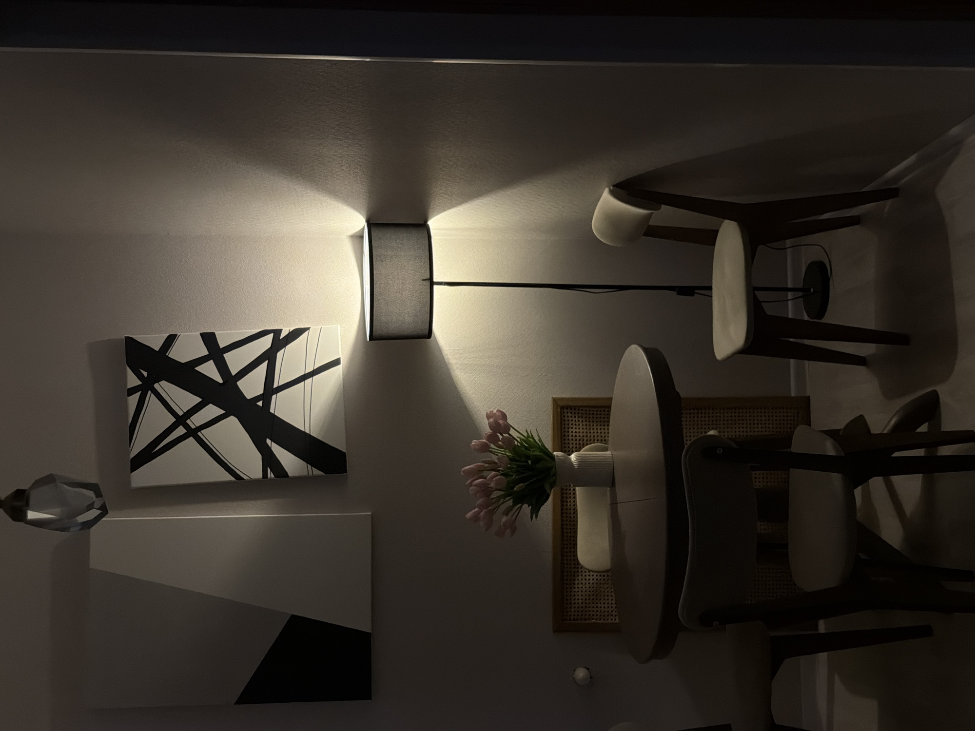 The image depicts an indoor scene, possibly a dining area, with warm lighting casting long shadows. On the wall, two abstract artworks are displayed: one with geometric shapes in black and white, and another with bold black brushstrokes on a white background. A floor lamp with a dark lampshade stands in the corner, its light illuminating the textured wall. In the center of the room, a round wooden table is set with a vase of pink tulips. Two chairs are positioned around the table, their seats appearing to be upholstered in a light-colored fabric. A woven rattan-style panel is visible behind the table, adding a natural texture to the scene. The overall ambiance is serene and somewhat dimly lit. No specific location in Moscow, Russia, is indicated by any visual cues in the image.