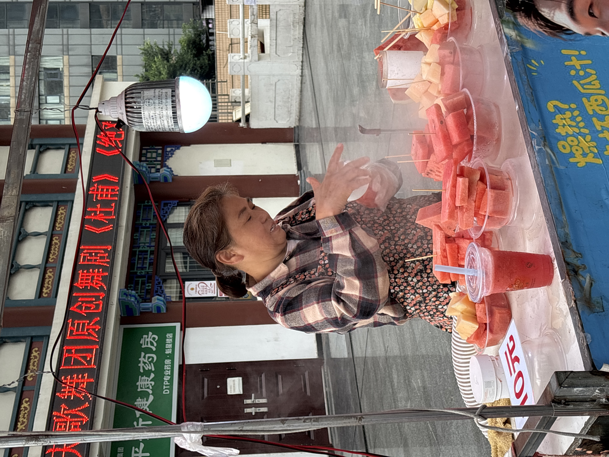 Here is a description of the image:
The image is an outdoor shot of a street scene in Yuzhong District, Chongqing, China. The main subject is a woman with her hair pulled back, wearing a plaid jacket over a floral dress. She is standing behind a makeshift fruit stand, which holds an array of cut fruits, including watermelon and melon, skewered on sticks or in plastic cups. A sign on the stand indicates a price of 10 yuan. The woman appears to be arranging or interacting with the fruit.
In the background, a building with ornate Chinese architectural details is visible. A red LED sign displays Chinese characters, with the text "大歌舞团原创舞剧《杜甫》集团" which translates to "Large Song and Dance Troupe Original Dance Drama 'Du Fu' Group." A green sign beside it reads "千于健康药房" (Qianyu Healthy Pharmacy) with "DTP专业药房·魁星楼店" (DTP Professional Pharmacy - Kui Xing Lou Branch) below it. An LED light fixture is attached to a pole above the woman's stand. Powerlines are running across the foreground. The surrounding environment suggests an urban street with pedestrian traffic.