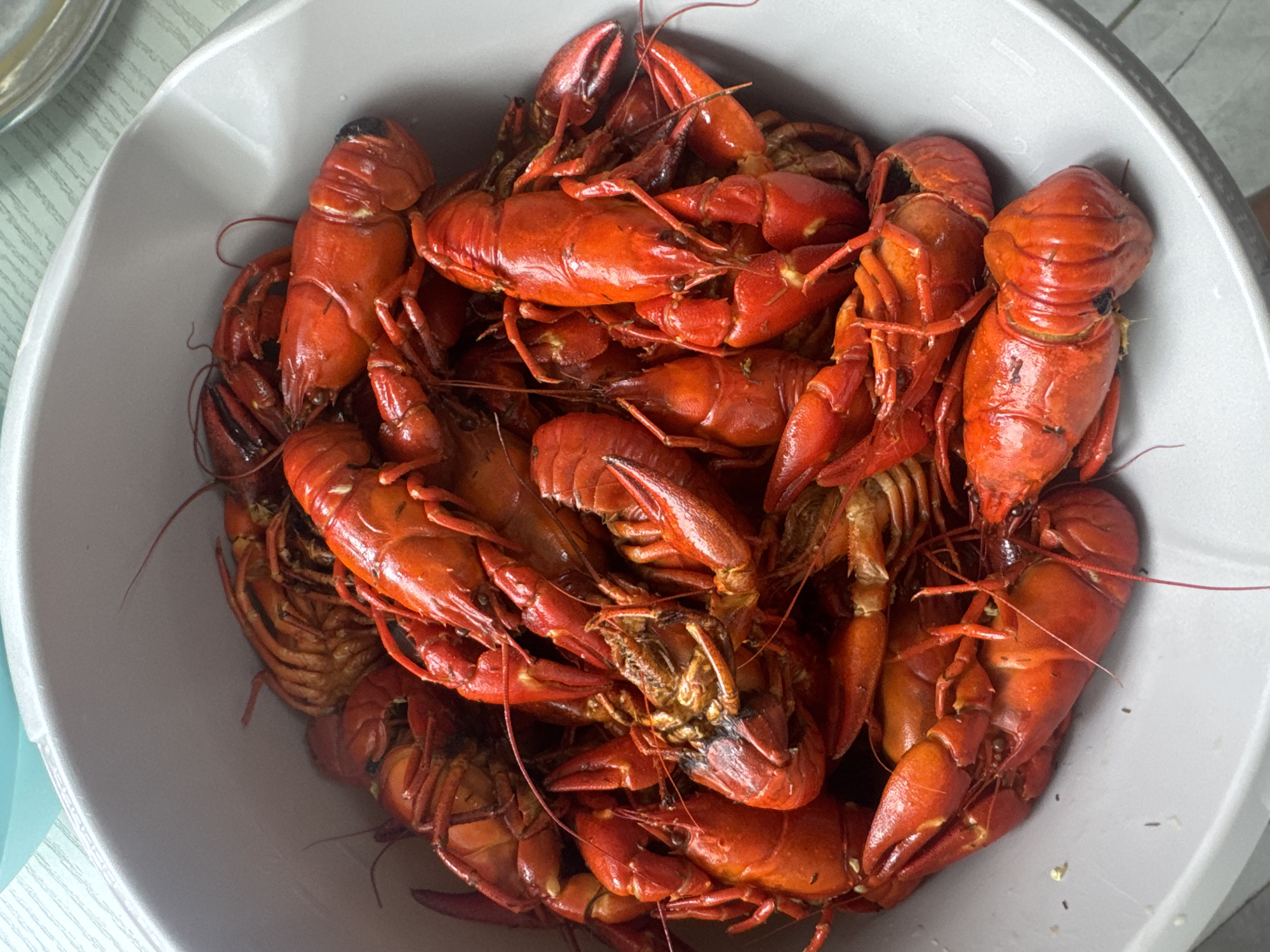 A close-up, overhead shot shows a large quantity of cooked crawfish piled high in a light gray bowl. The crawfish are bright red and have a textured shell. Some crawfish are whole, while others appear to have been separated. The bowl is positioned on a surface with a light green and white striped pattern, possibly a tablecloth or placemat. The background is blurred, with a hint of a light blue object in the upper left corner and a silver bowl in the upper right. The image is well-lit, emphasizing the vibrant color of the crawfish. No specific location in Duren, Germany, can be determined from the visual information provided.