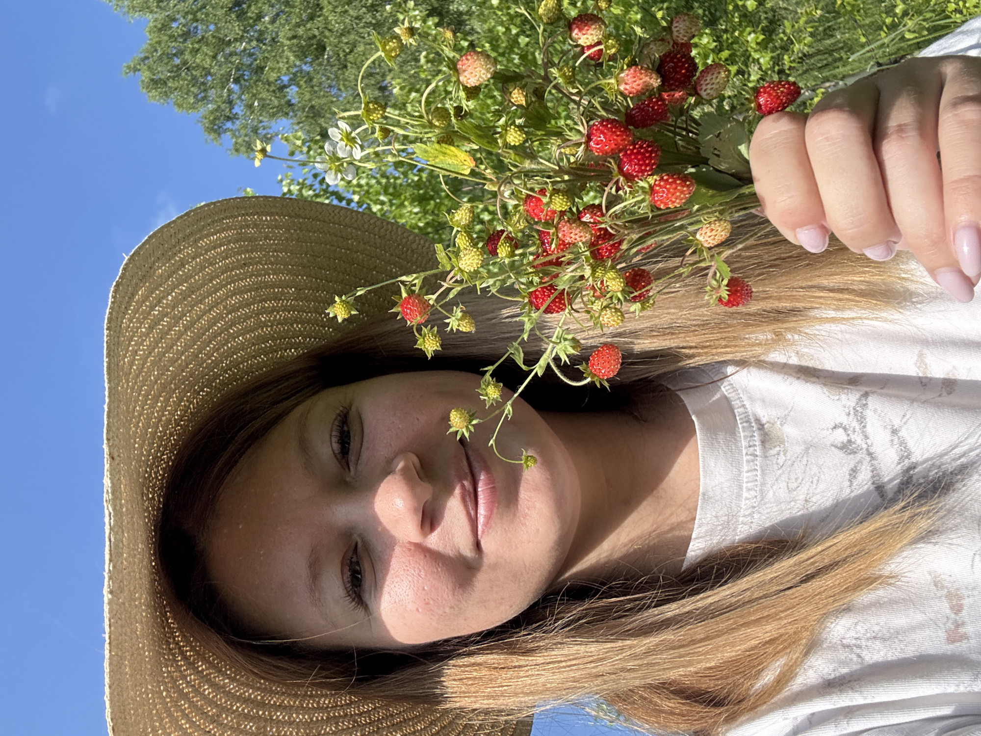 A medium close-up selfie shows a young woman holding a bouquet of wild strawberries. She is wearing a straw hat and a white t-shirt with a faded pattern. The background is a bright blue sky and lush green trees. The woman is smiling and looking at the camera. The wild strawberries are a mix of red and green, with some white flowers. The image appears to have been taken outdoors on a sunny day. There is no indication of the location in Vassyata, Russia.