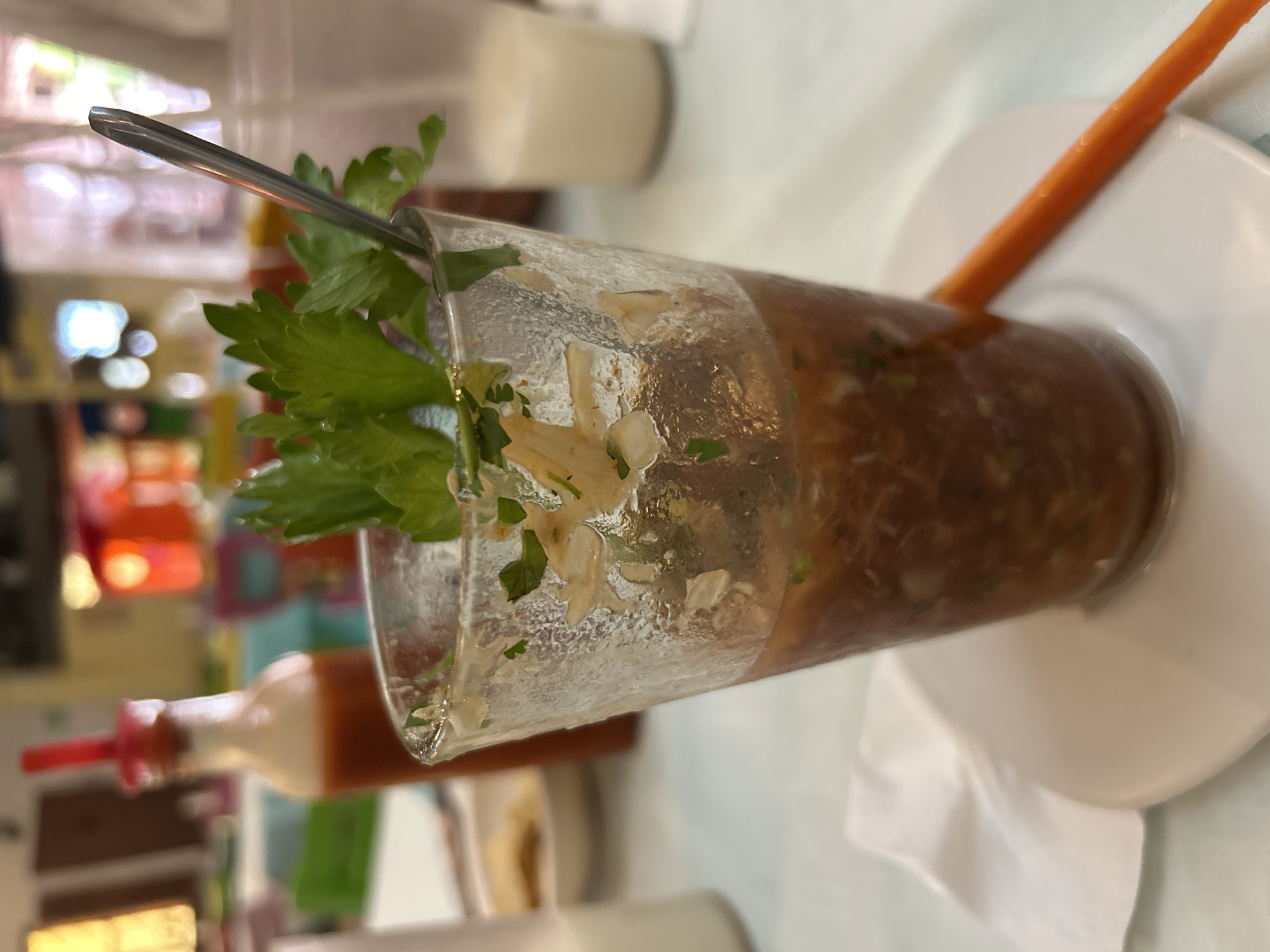 The image captures a prawn cocktail in a glass, garnished with celery. A spoon is seen in the glass. The drink rests on a white plate, alongside a carrot stick. In the background, a bottle of sauce is visible. The location is Veracruz City, Mexico, judging by the presence of the prawn cocktail. The scene appears to be indoors, possibly a restaurant.