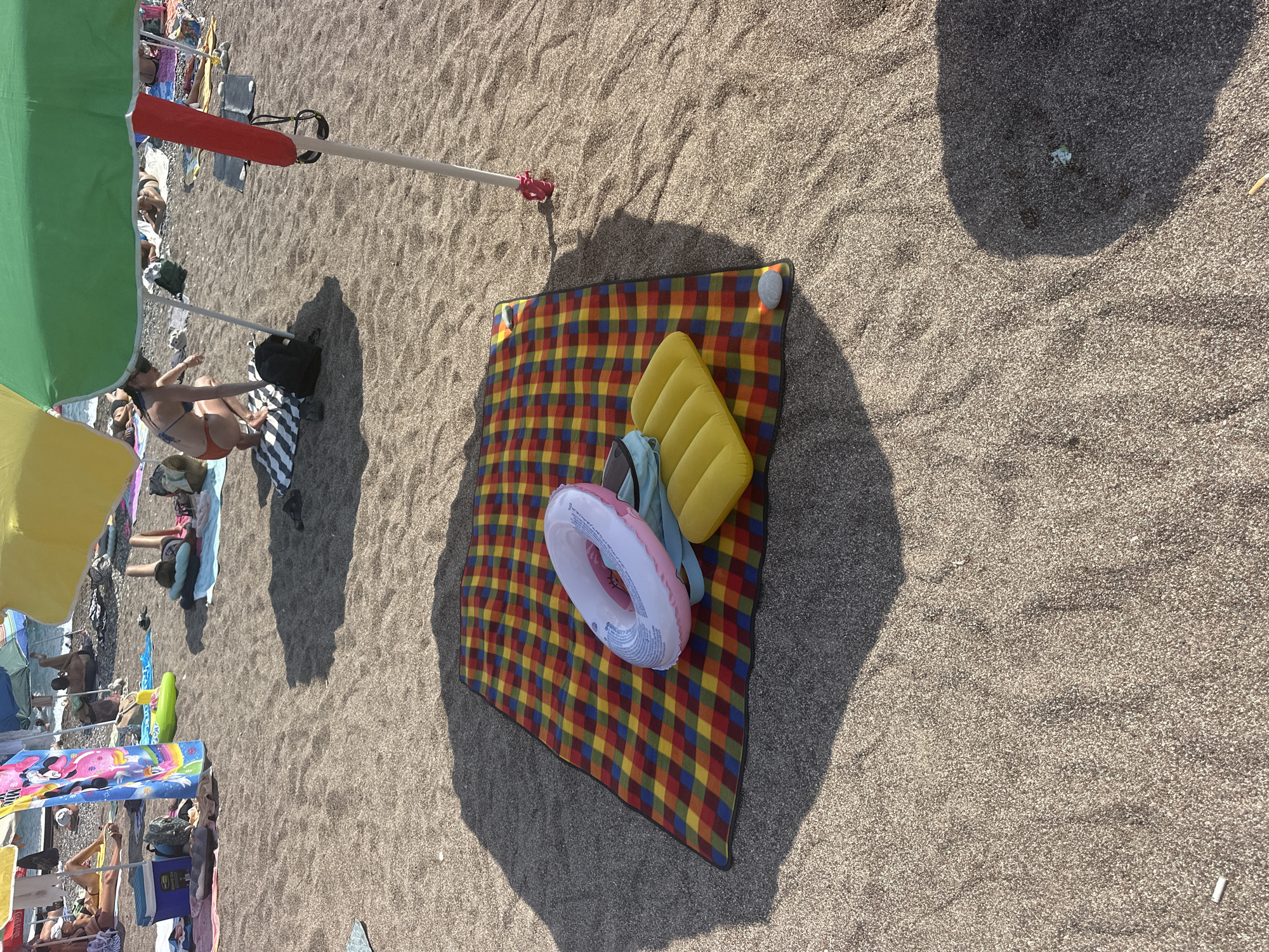 The image shows a beach scene in Boreti, Montenegro, with several people relaxing on the sand. In the foreground, a colorful plaid blanket is spread out, with an inflatable ring and a yellow inflatable mat placed on top. A beach umbrella casts a shadow over the blanket. In the background, other beachgoers are seen lying on towels or sitting, with more umbrellas providing shade. The sea is visible in the distance. The sand appears to be dark and coarse, typical of a pebble beach.