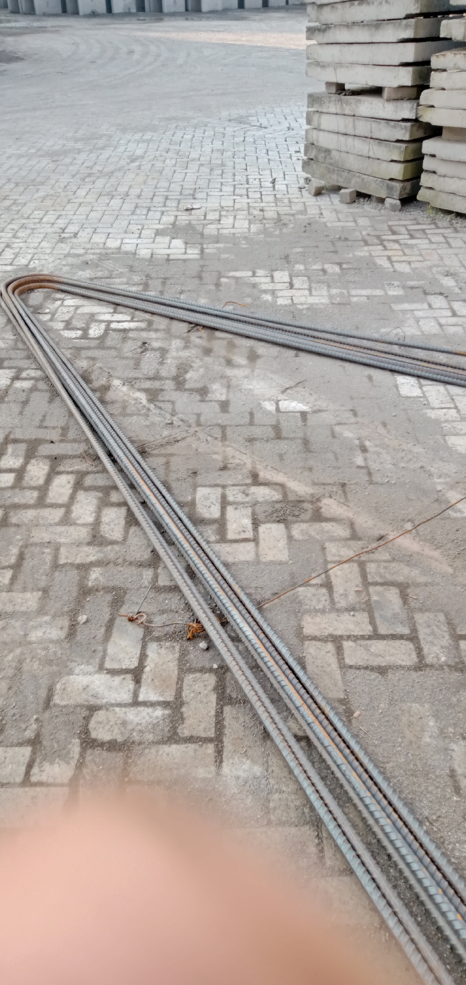 The image depicts a set of reinforcement steel bars, also known as rebar, laid out on a paved surface in Sumput, Indonesia. The rebar appears to be new and unused, characterized by a ridged texture. The bars are configured in a partially rectangular form with rounded corners. The paving material consists of light-colored brick laid in a pattern. The background shows stacked, concrete slabs. The lighting indicates it's daytime. A blurry human hand is intruding from the lower-left of the image.