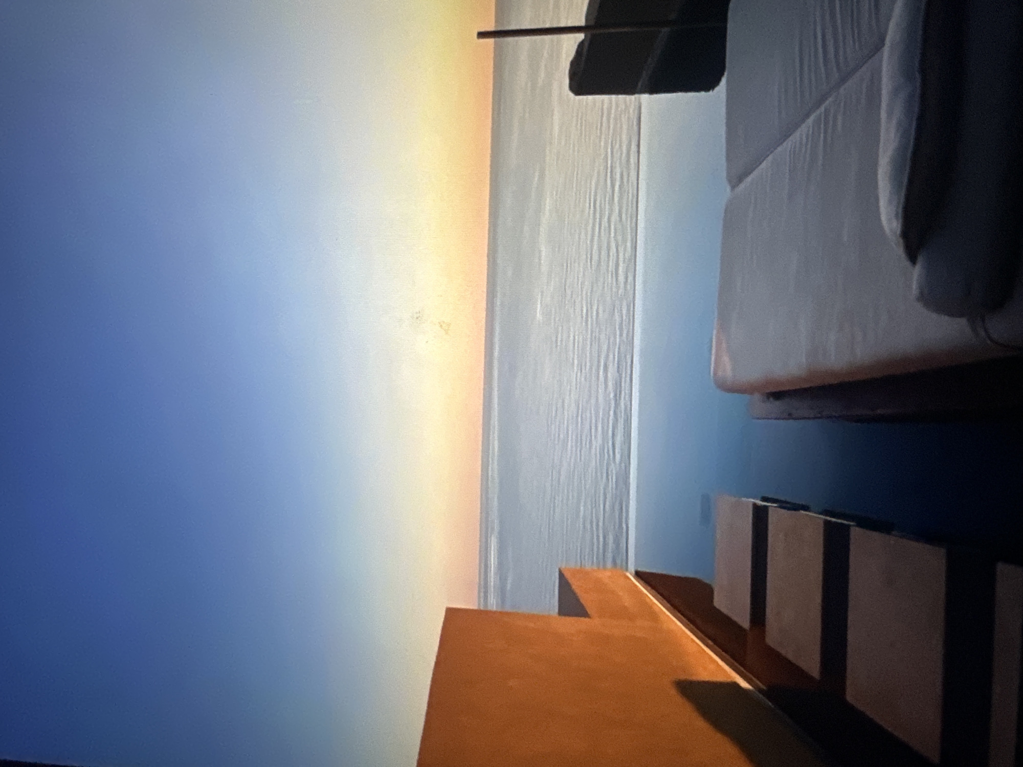 An outdoor infinity pool overlooks the ocean during a serene sunset. In the foreground, two lounge chairs are positioned side-by-side. To the left, a large, solid orange structure juts out into the frame, casting a shadow onto a series of flat, square stepping stones that lead into the dark water of the pool. The ocean stretches out to the horizon, with a clear sky above transitioning from a deep blue at the top to a soft gradient of yellow and orange near the horizon, indicating the sun is setting.