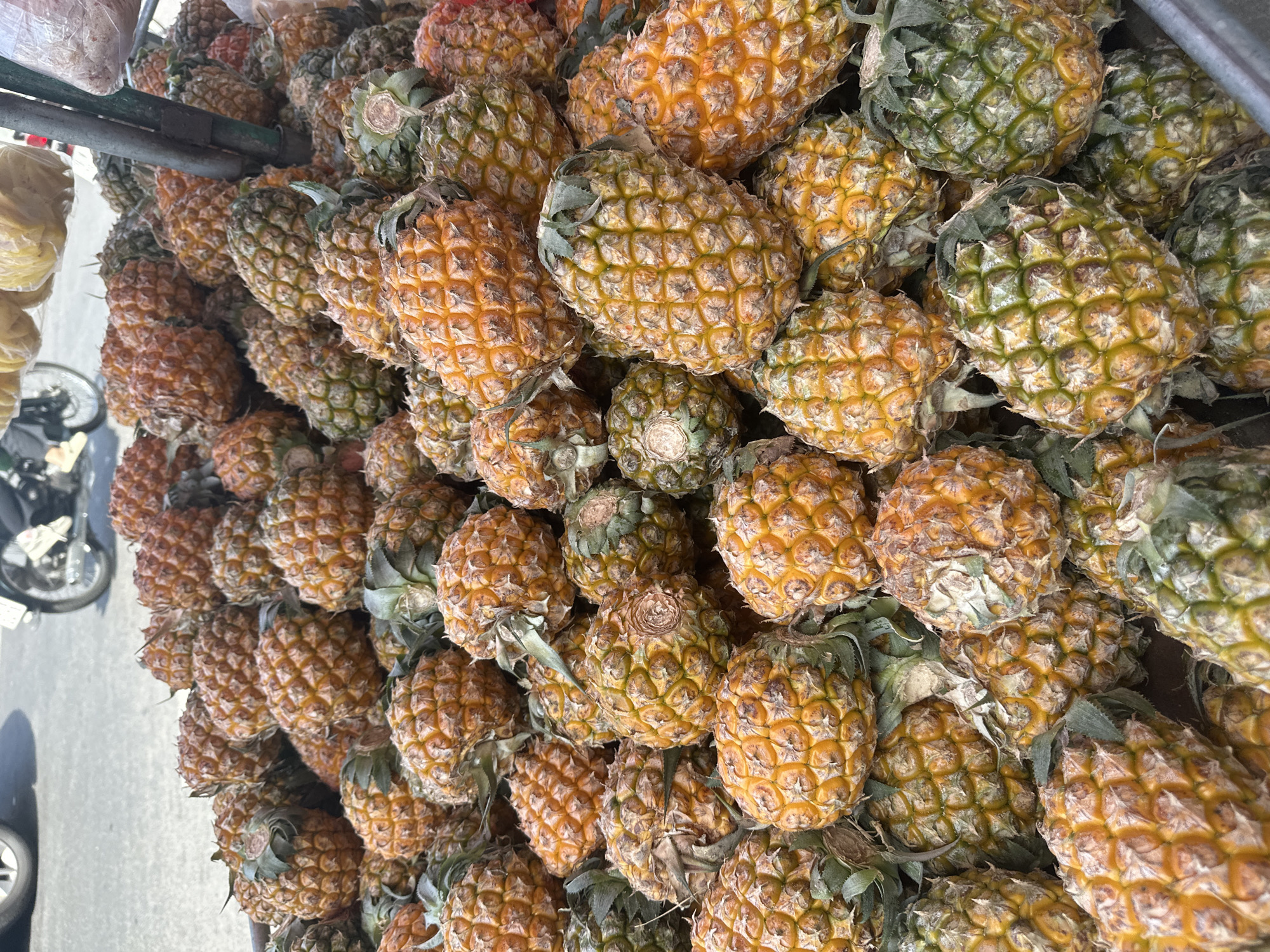 A large pile of ripe pineapples is displayed on a street stall in Bac Nha Trang Ward, Vietnam. The pineapples are a mix of yellow and green, with their spiky skin clearly visible. Some have green leafy tops. The stall appears to be on a paved street, with a motorcycle and its rider visible in the blurred background to the upper left. There are also other items on the stall, possibly more fruit, partially obscured by a metal pole in the center right of the image. The scene suggests a typical outdoor market in Vietnam.