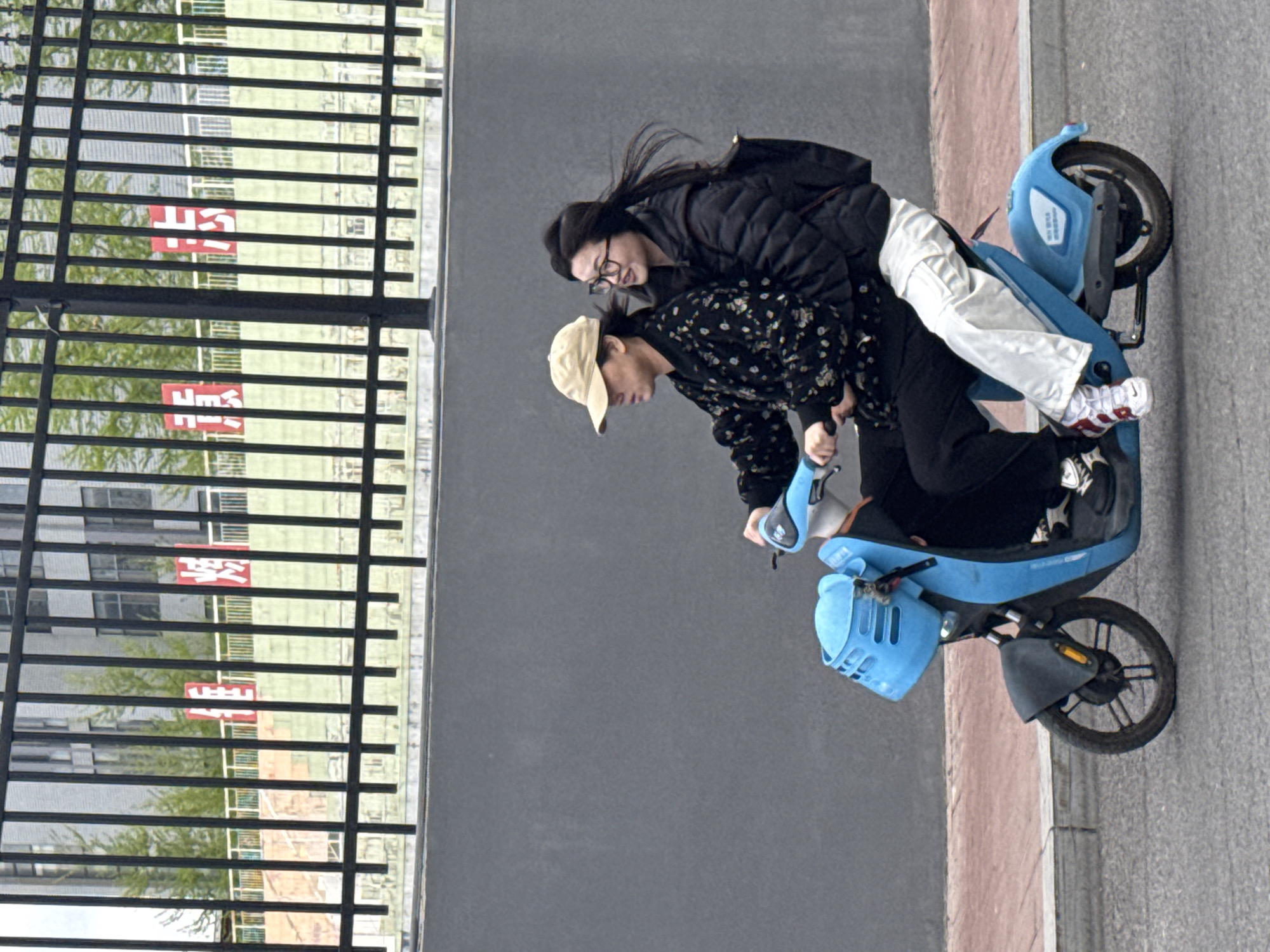 The image, set in Zhongtun, China, depicts two individuals on a light blue electric scooter on a paved road or sidewalk. The scene suggests an urban or campus environment, viewed from a slightly elevated perspective.
The person in the front, appearing to be the driver, wears a light-colored baseball cap, a dark long-sleeved top with a subtle pattern, and dark pants. They are focused downwards, holding the handlebars of the scooter. Behind them, a passenger with glasses, a black puffer jacket, white pants, and red-and-white sneakers stands on the scooter's footrest, holding onto the driver. This individual looks towards the camera with a gentle smile.
The electric scooter is prominently light blue, featuring a front basket and standard handlebars. To the left, a black metal fence or railing runs horizontally across the frame. Beyond the fence, a verdant area of trees or bushes precedes multi-story buildings, which have numerous windows.
Visible through the fence are two red rectangular signs. Closer inspection reveals Chinese characters on these signs. The characters "学" (xué - study) and "中" (zhōng - middle) are discernible, suggesting the term "中学" (zhōngxué - middle school), indicating the background is likely a school campus. The lighting is soft and even, characteristic of daytime, possibly an overcast day, and the mood appears casual and relaxed.