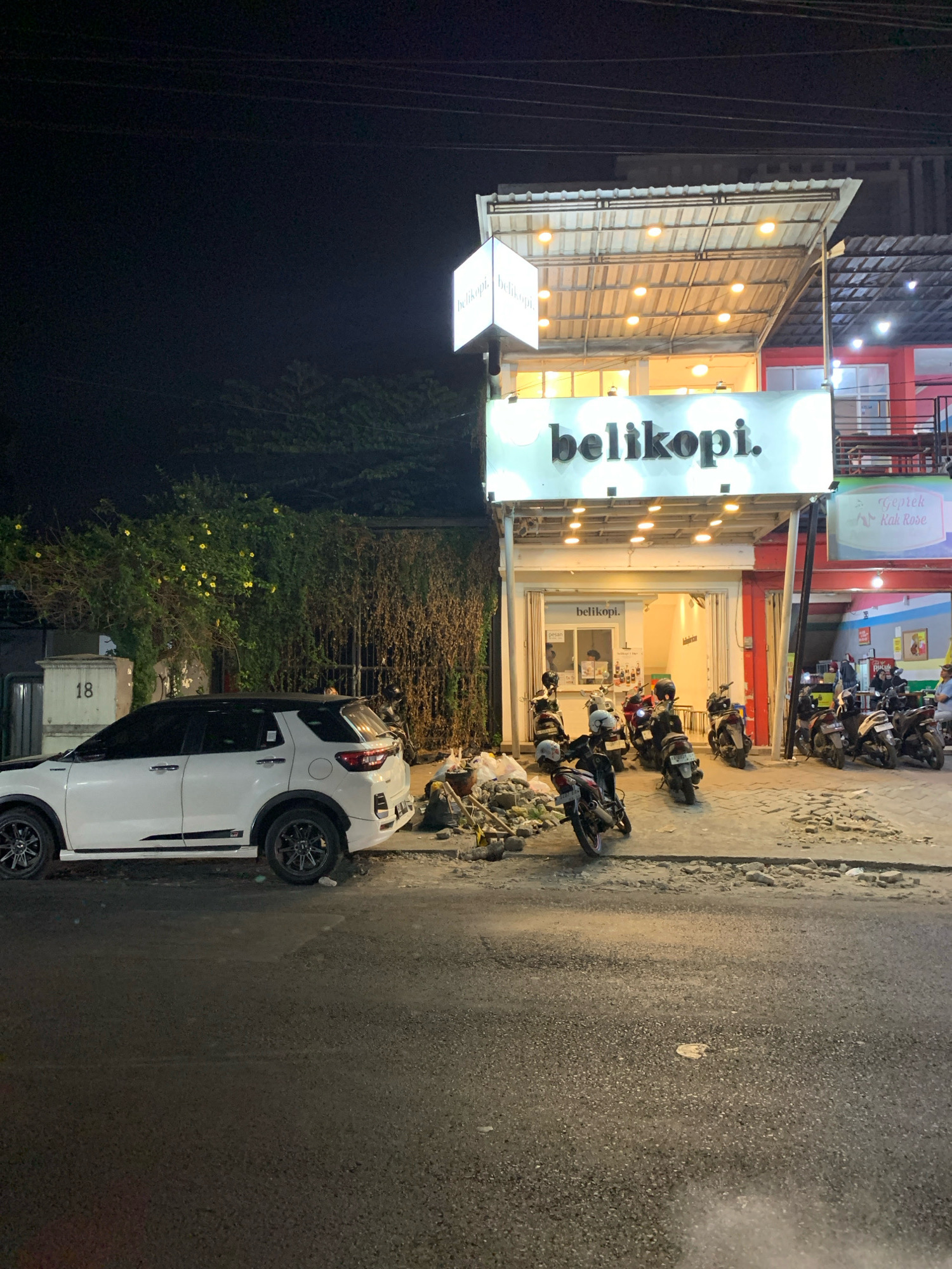 At night, a white SUV is parked on the side of the road in front of a building with an illuminated sign that reads "belikopi." The building appears to be a coffee shop or cafe, with a covered outdoor seating area and glass doors. Several motorcycles are parked in front of the building. The street in front of the building is paved, and there is a patch of dirt and debris to the left of the building. The area appears to be a commercial district, with other buildings and businesses visible in the background. The number "18" is visible on a wall to the left of the SUV, which may indicate a street address. The overall scene suggests a lively nighttime atmosphere in a city.