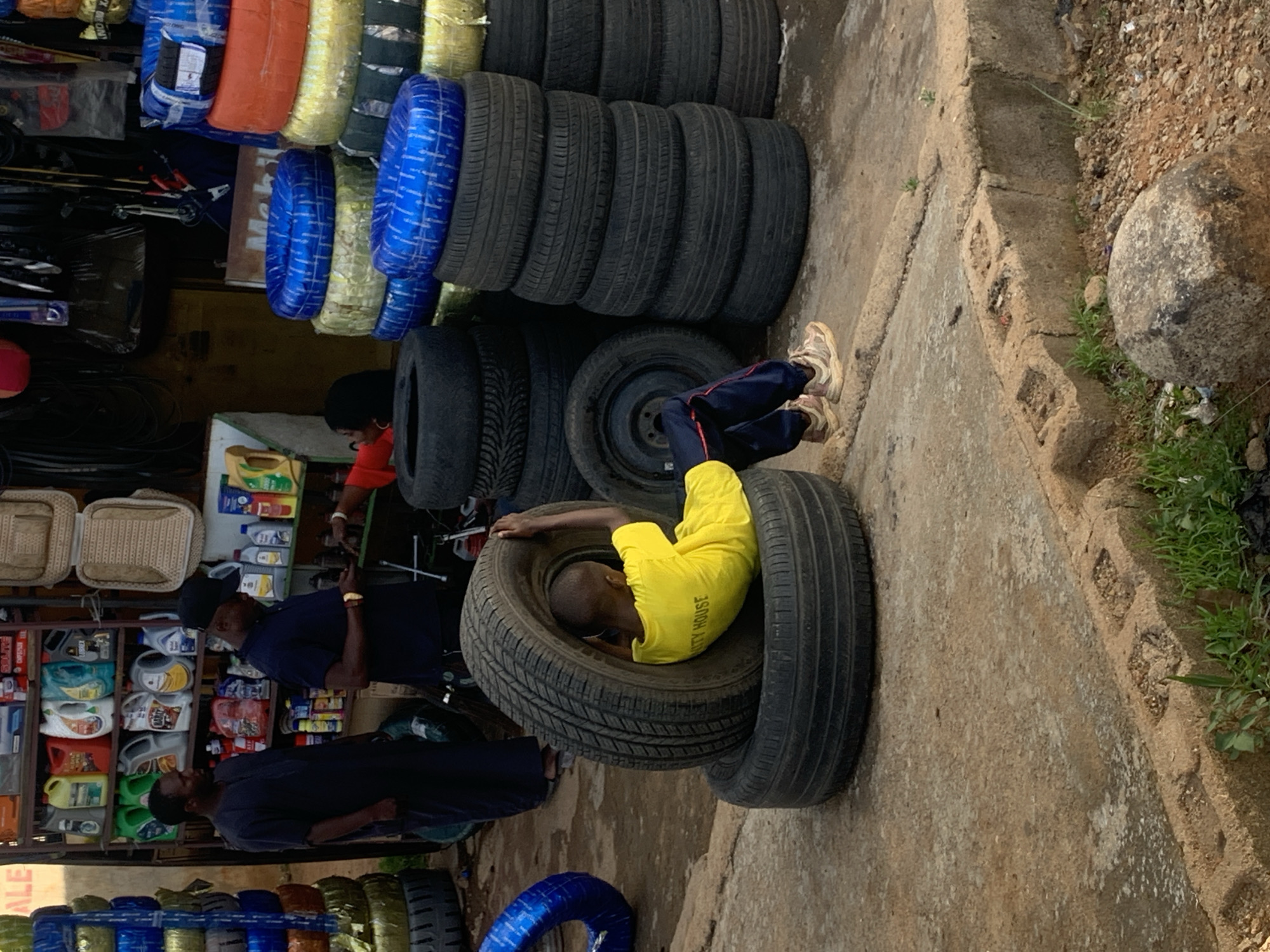 The image depicts a scene outside a shop in Jos, Nigeria, likely a tire or auto parts store given the stacks of tires visible. In the foreground, a young boy wearing a bright yellow t-shirt and dark pants is sitting inside a large tire, his legs extended. He is positioned within two stacked tires. Behind him, two men are standing. One man, dressed in a dark robe, faces away from the camera and appears to be looking at the shelves of products in the shop. The second man, wearing a dark t-shirt and a black baseball cap, is also facing the shop interior and appears to be looking at his phone. The shop itself has shelves stocked with various items, including bottles of liquids, likely automotive fluids, and other auto parts. Large stacks of new tires, some wrapped in blue plastic, are piled up outside the shop. The ground is a mix of concrete and dirt, with some sparse vegetation and a large rock visible in the lower right corner. The overall atmosphere suggests a commercial area in an urban setting.