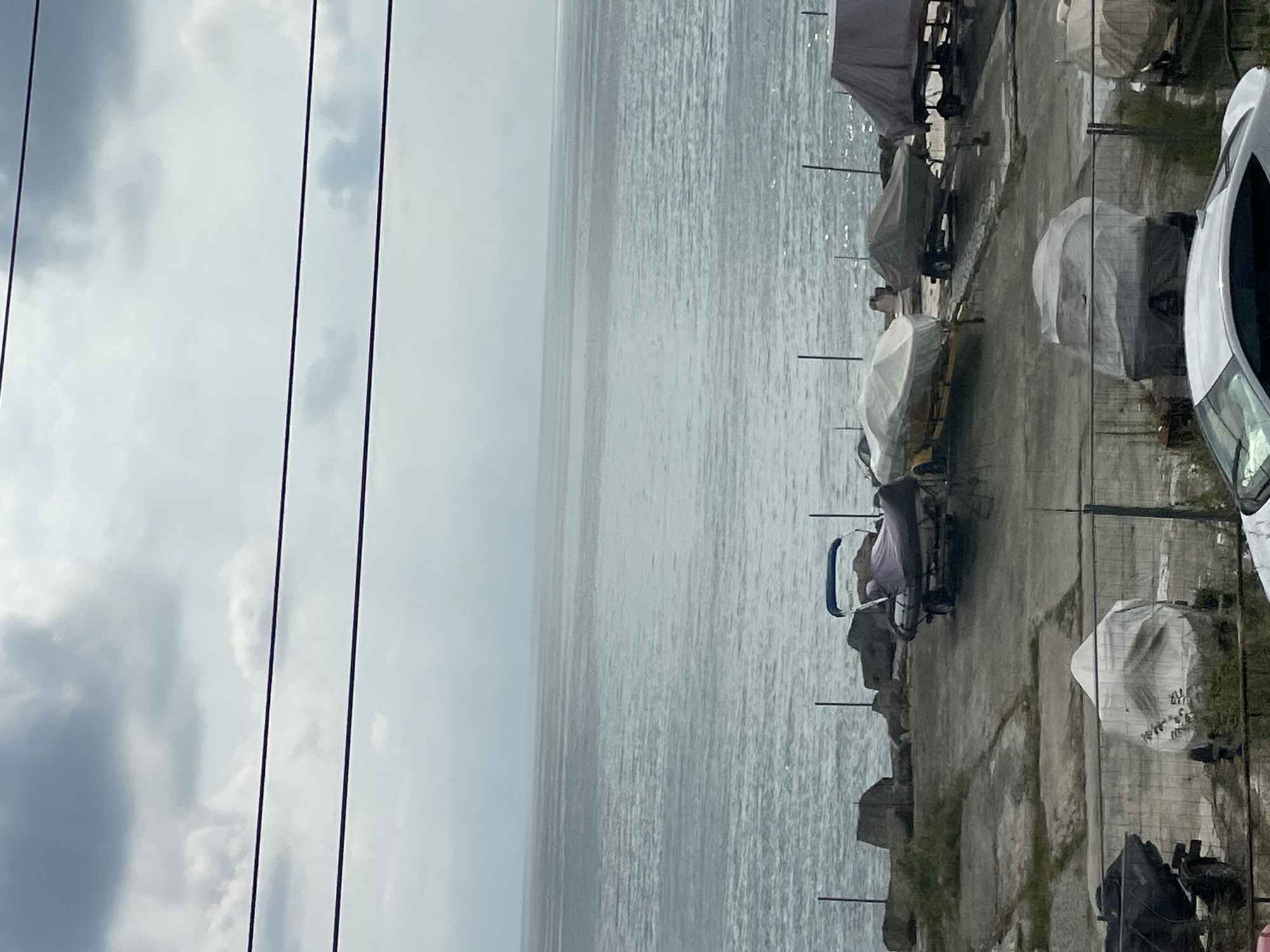 The image depicts a view of the Black Sea in Sochi, Russia, from what appears to be a coastal area with boats. The sky is overcast with gray clouds, and the sea is calm with gentle waves. In the foreground, several boats are parked on a paved area, some covered with tarpaulins. A few of the boats are on trailers. There is also a fence in the foreground, and a white car is partially visible. The general atmosphere suggests a quiet, possibly off-season, scene at a marina or boat storage area.