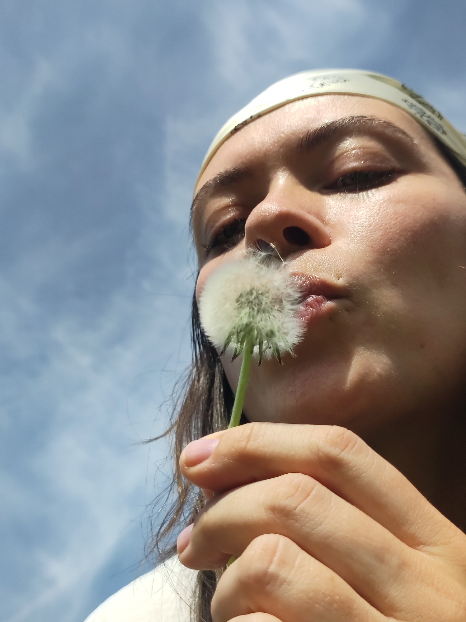 A low-angle, close-up shot depicts a woman with fair skin blowing on a dandelion. She is wearing a patterned bandana. Her dark hair is visible along her shoulders. The background is a blue sky with scattered white clouds. The location is not discernible from the image.