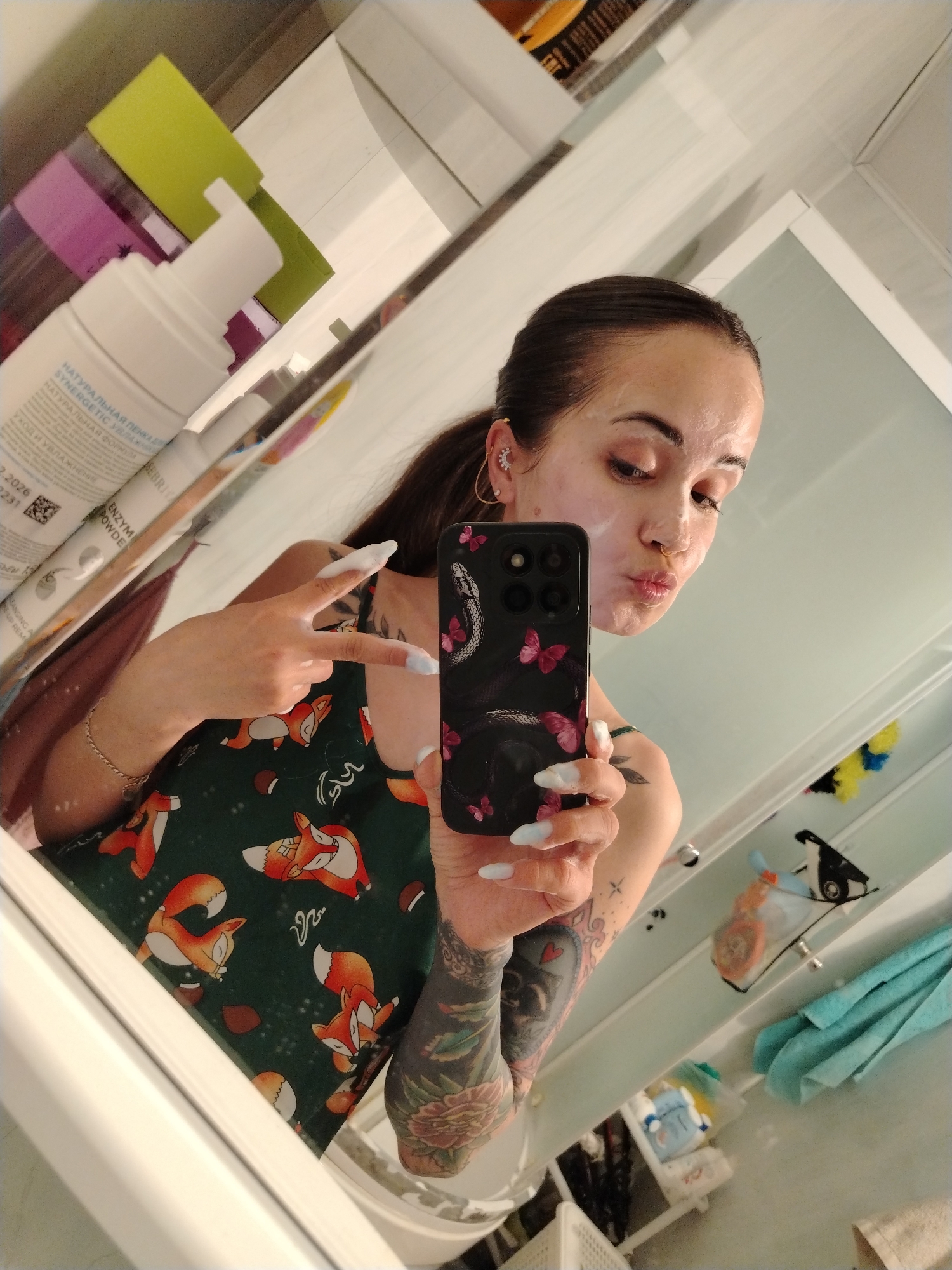 A young woman is depicted taking a mirror selfie in a bathroom, likely in Noviy, Russia. She has a white mask or skincare product applied to her face, partially covering her eyebrows, nose, and chin. Her expression is playful, with puckered lips. She is wearing a dark green top featuring an orange and white fox and acorn pattern. Her left arm, heavily adorned with dark floral tattoos, is visible in the reflection. She also has a nose ring and a moon-shaped earring on her right ear.
She holds a smartphone with a black case decorated with a snake and pink butterflies. Her long, light blue artificial nails are prominent, with some of the white face product visible on her fingers. In one instance, she makes a peace sign with her right hand.
The bathroom setting is evident from the white tiled walls, a shower enclosure, and various reflected toiletries. A white pump bottle with Cyrillic text is clearly visible, showing phrases like "НАТУРАЛЬНАЯ ПЕНКА ДЕ" (Natural foam for), "SYNERGETIC", "НАТУРАЛЬНАЯ ФОР" (Natural form), "УХОДИ УВЛАЖНЕНИЕ" (Care and hydration), and "ENZYM POWDE". Other details such as "BERRIT", "2.2026", and "2231" are also legible. A bright green sponge and other bathroom items can be seen in the background. The scene suggests a casual, self-care routine, likely in the evening.